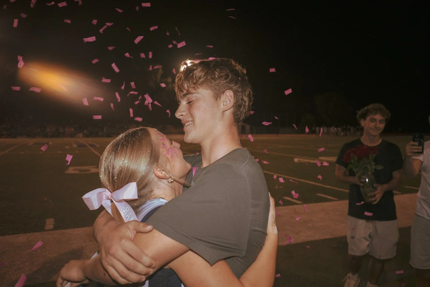 Two teens hug as pink confetti falls on a football field, with stadium-light flare glowing in the corner