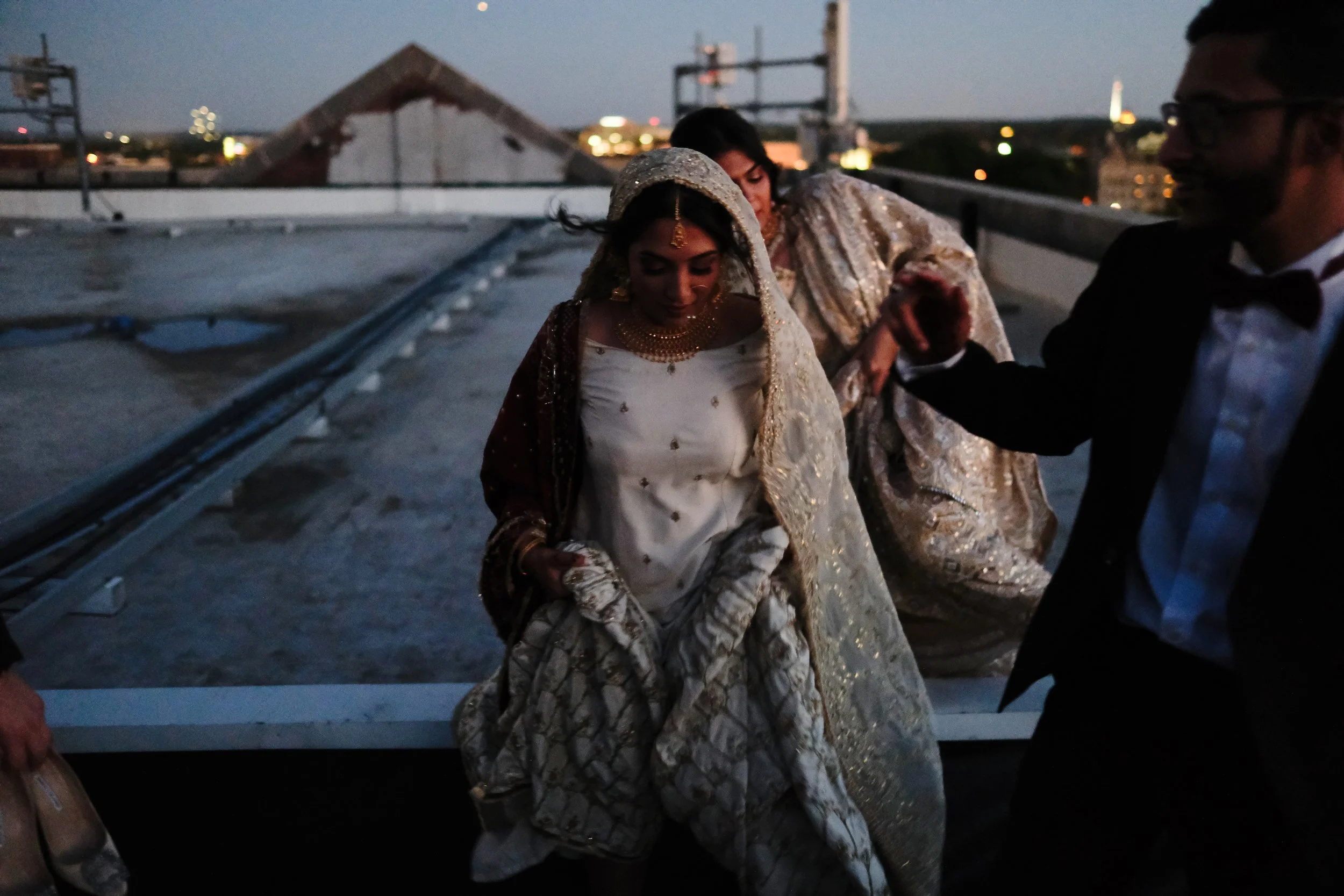 A bride in traditional Indian wedding attire, wearing a cream and gold saree with heavy jewelry and a veil, on a rooftop during evening, with a man in a tuxedo near her and another woman assisting her.