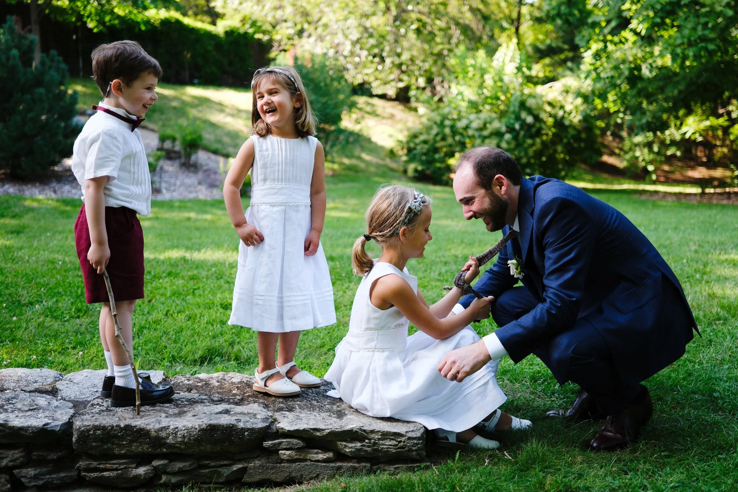 A man in a navy suit kneels on a grassy lawn, holding the hands of a girl in a white dress who is sitting on a stone wall, while three children in white and maroon outfits stand nearby, all laughing and enjoying an outdoor gathering in a lush garden.