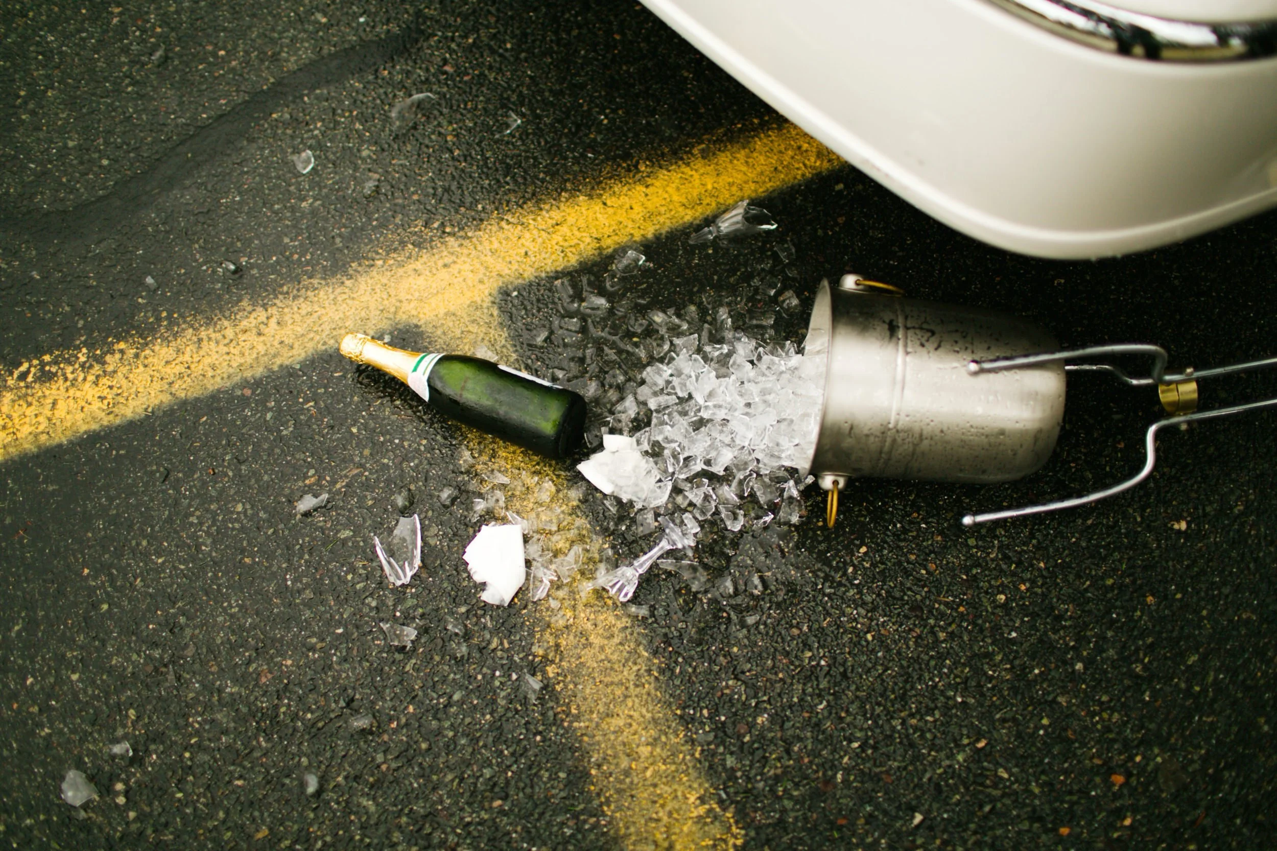 A champagne bottle and an exploded champagne corkscrew on the ground of a parking lot, with shattered glass and debris around.