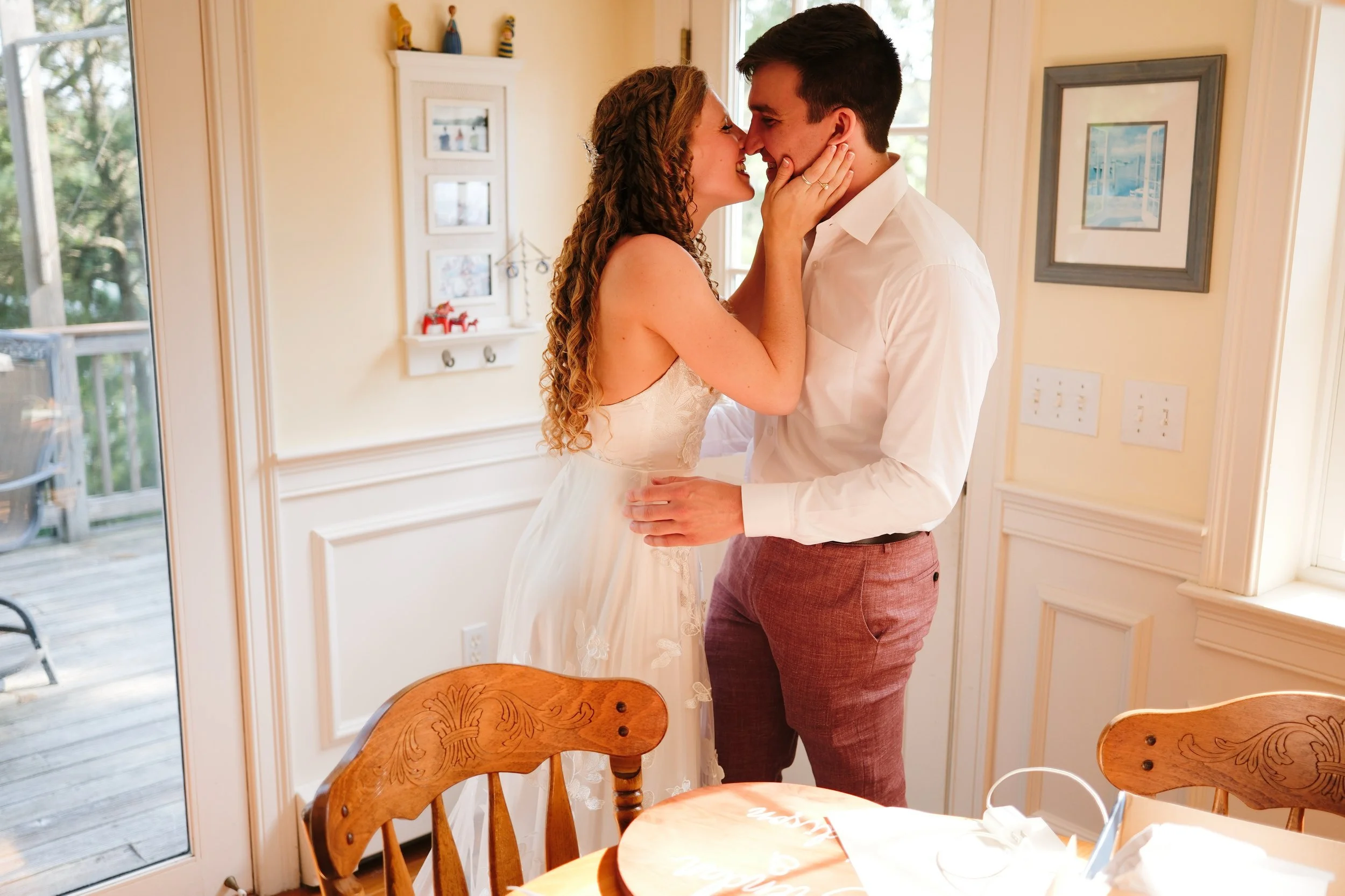 A woman and a man are embracing and touching noses indoors near a window and a door, with the woman holding the man's face and smiling.