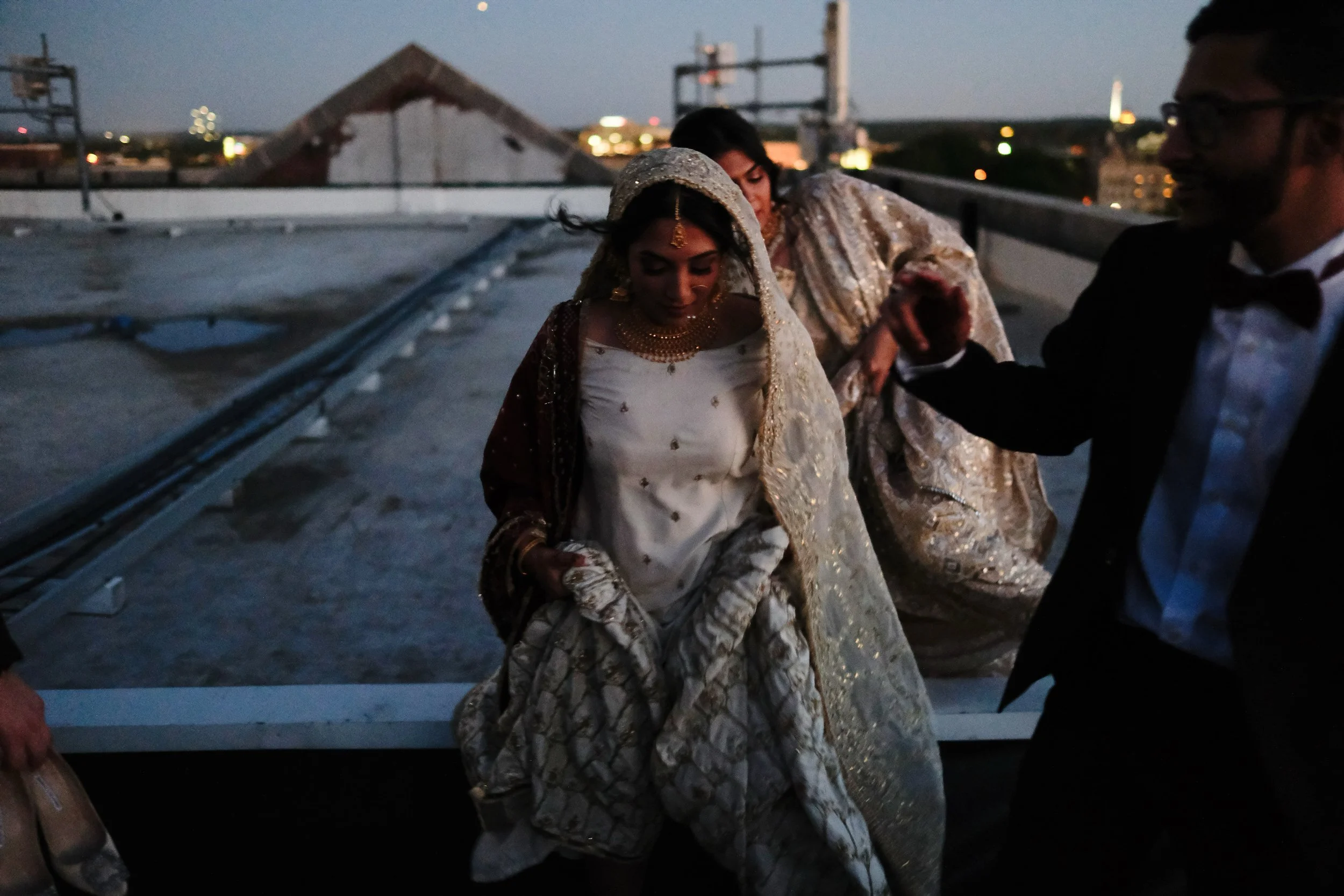 Indian bride in traditional wedding attire descending from a rooftop. She is wearing a white and gold embroidered saree with jewelry, accompanied by another woman and a man in formal wear.
