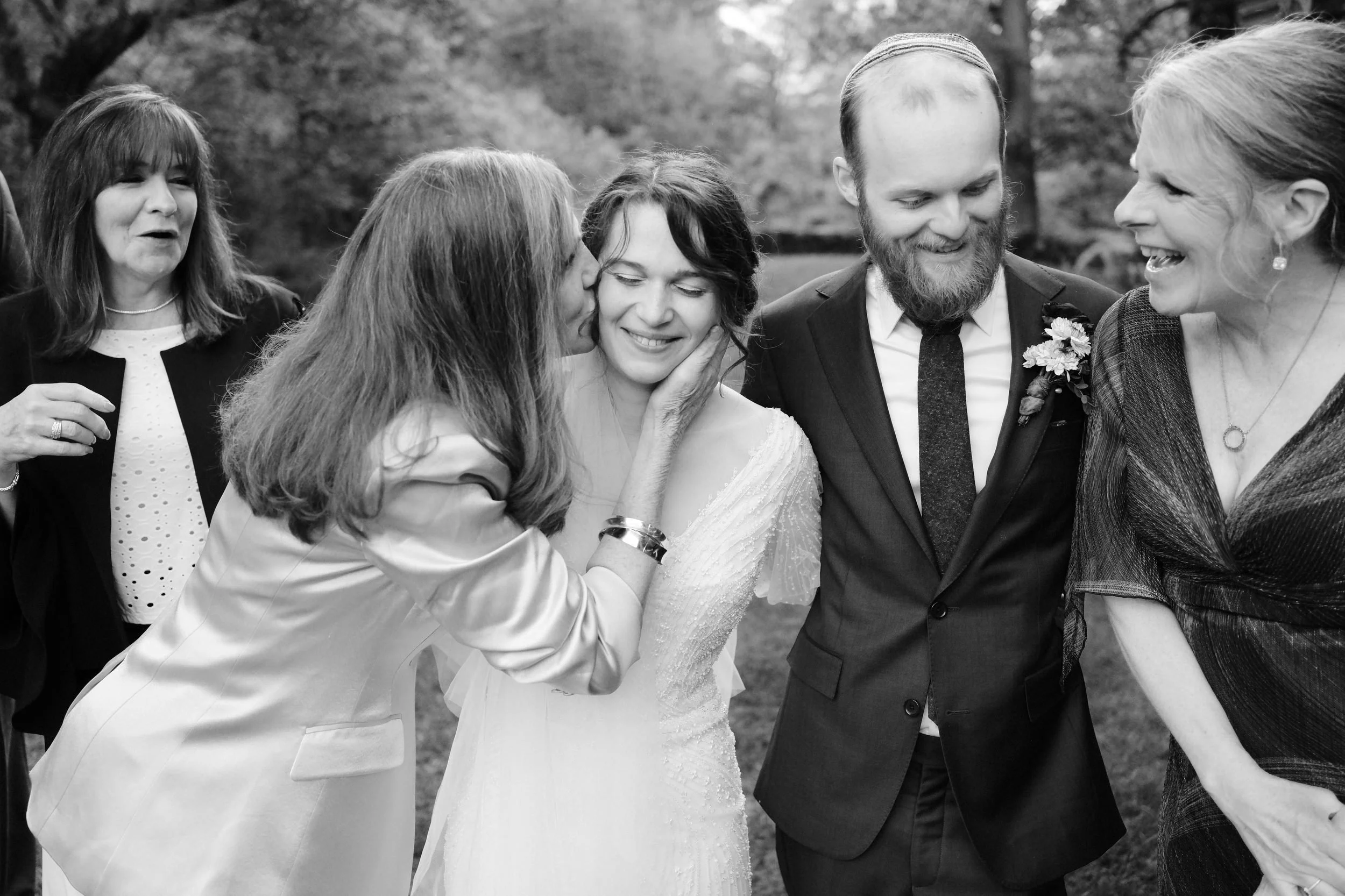 Group of people at a wedding, with two women kissing the bride on the cheek, and others smiling and laughing outdoors.