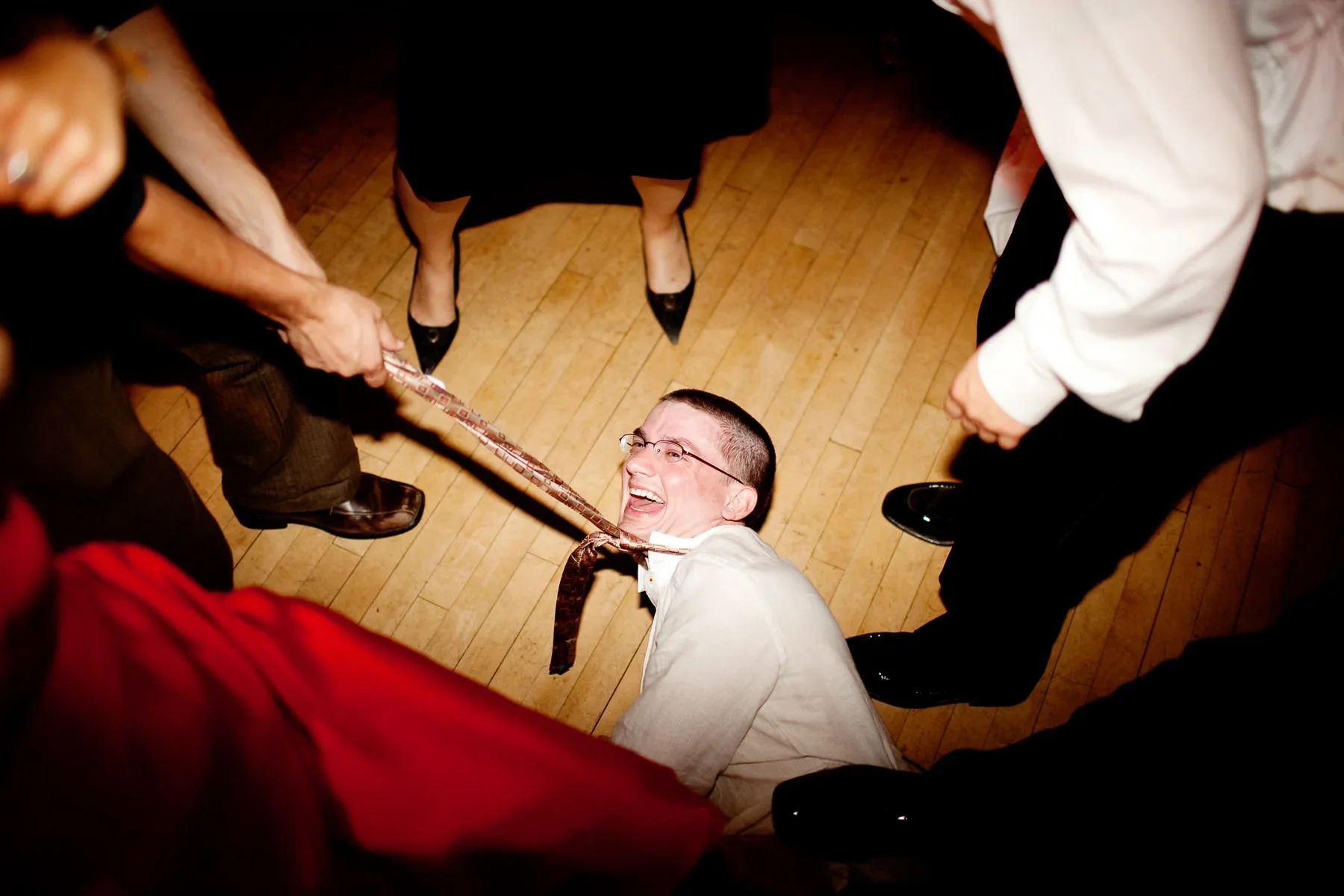 Man lying on the floor laughing as a woman pulls a tie around his neck at an event, surrounded by other people.