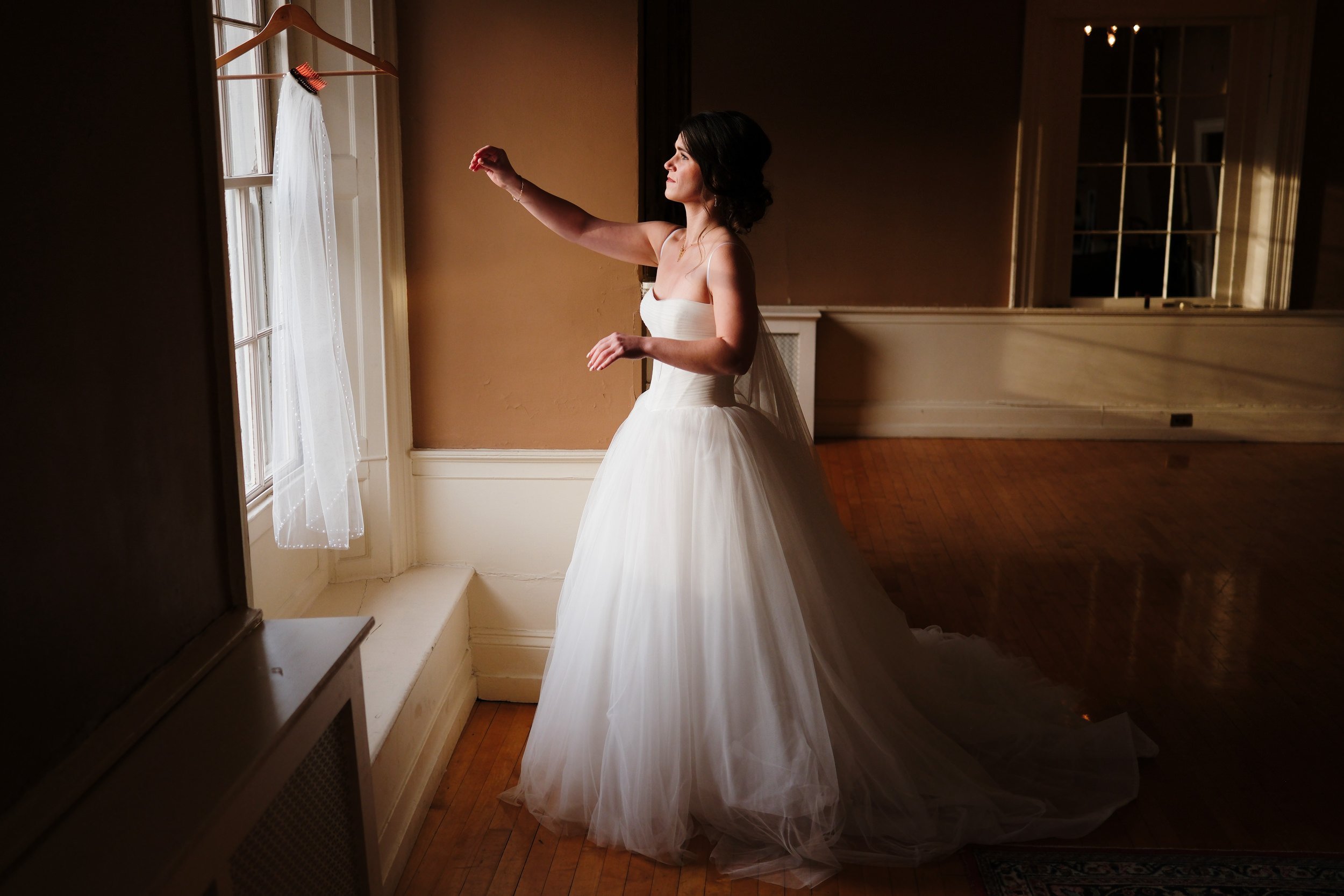 A bride in a white wedding dress standing by a window with sheer curtains, looking outside, with sunlight illuminating her and the room.