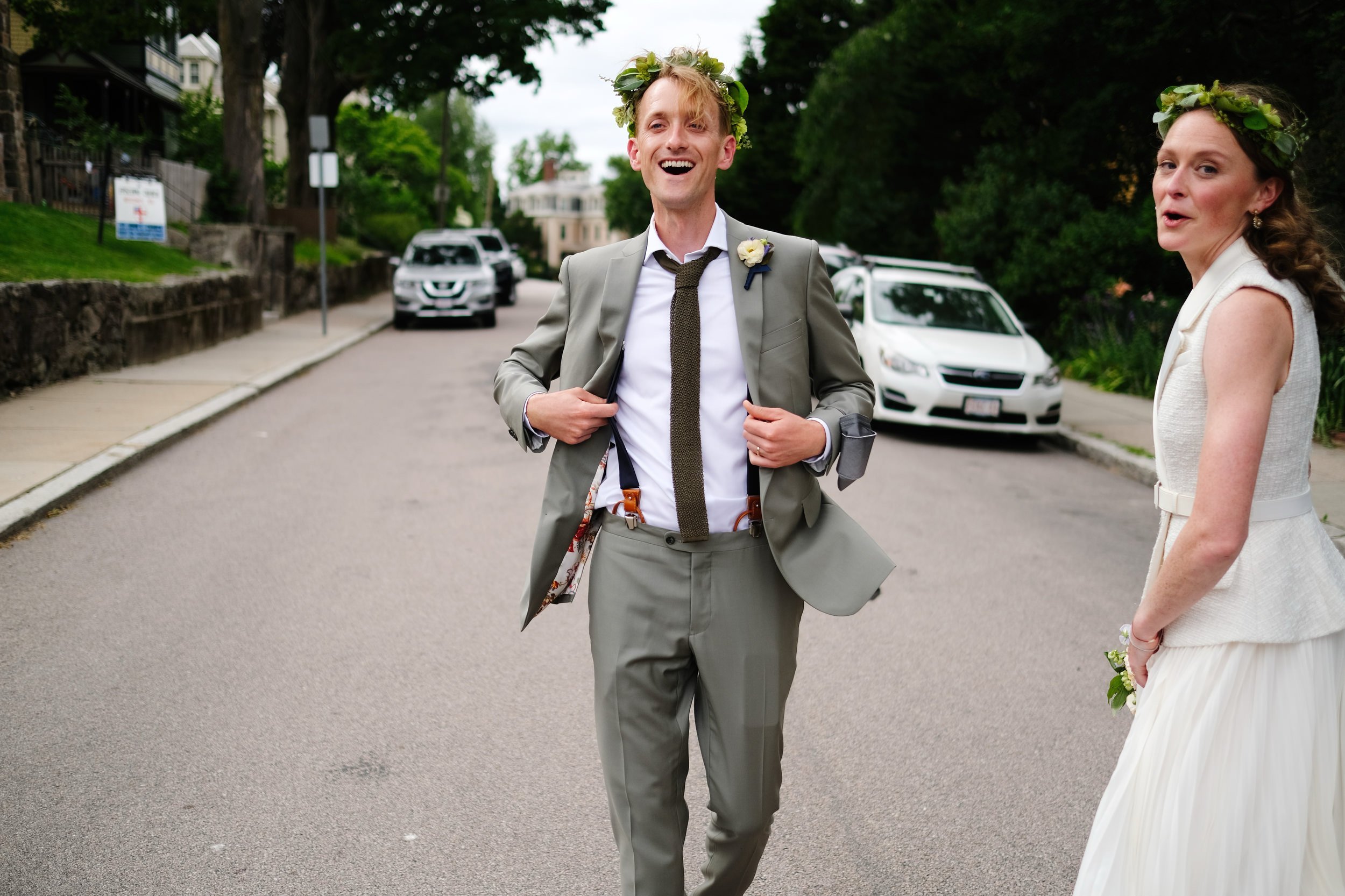 Happy man in a suit and woman in a white wedding dress standing on a residential street with cars parked behind them. Both are wearing flower crowns.