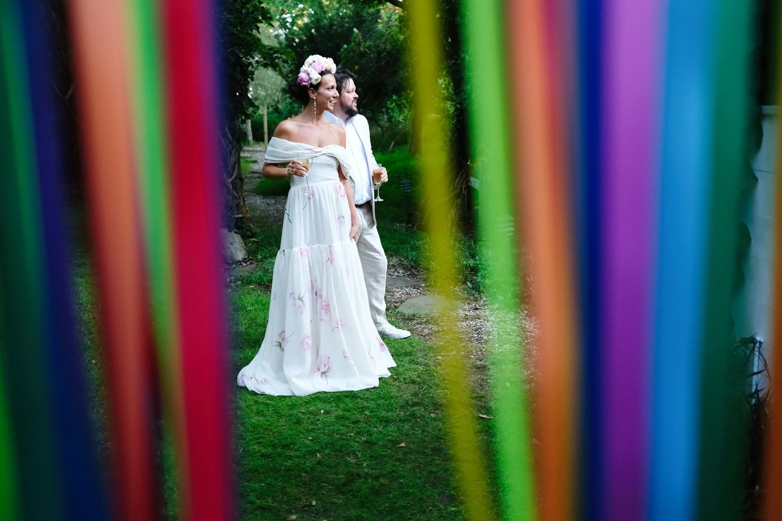 A woman in a white floral dress and floral headpiece and a man in a white suit and shirt are standing together outdoors, holding champagne glasses, seen through colorful vertical ribbons.