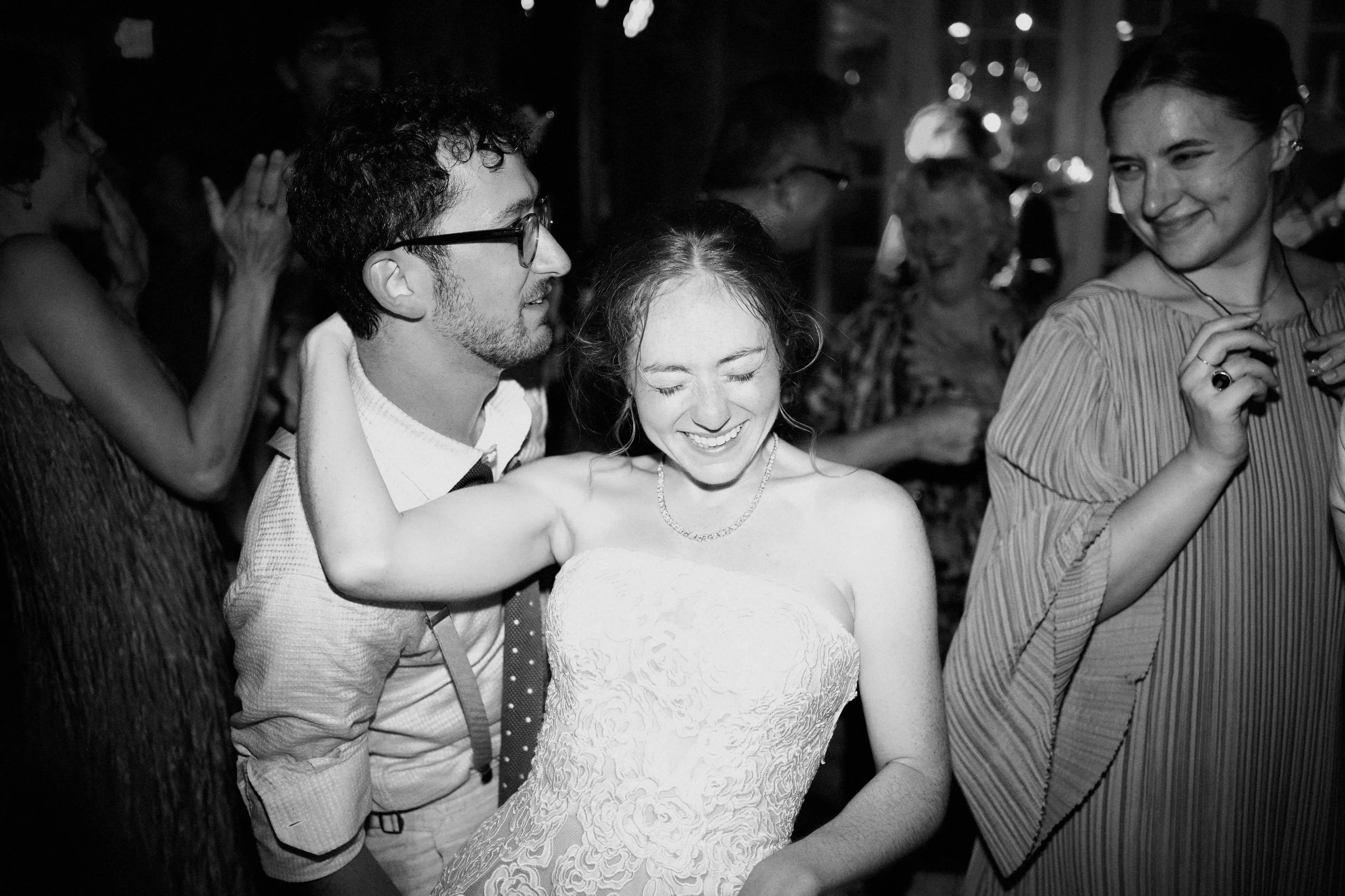 A black-and-white photo of a joyful wedding reception scene with a bride in a strapless lace wedding dress dancing and smiling, surrounded by guests, including a man with glasses and a woman holding a drink.