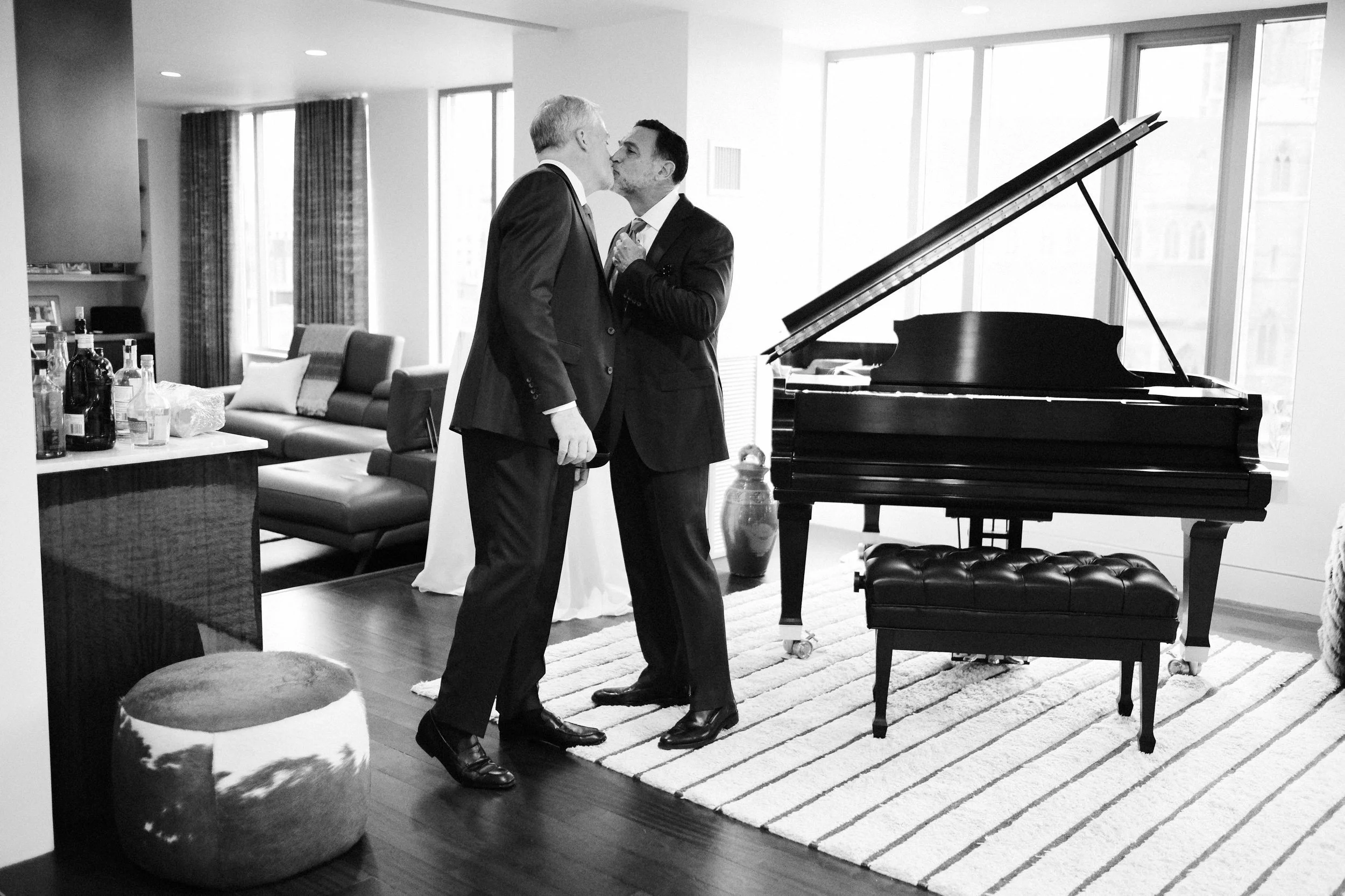 Two men in suits sharing a kiss in a living room with a grand piano and large windows.