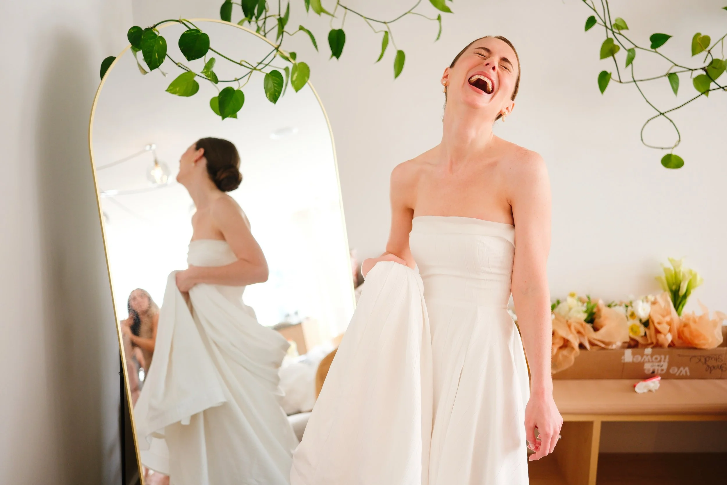 A woman in a white strapless dress, laughing joyfully while holding the side of her dress, standing in a room with a large mirror reflecting her back and a nearby table with flowers.