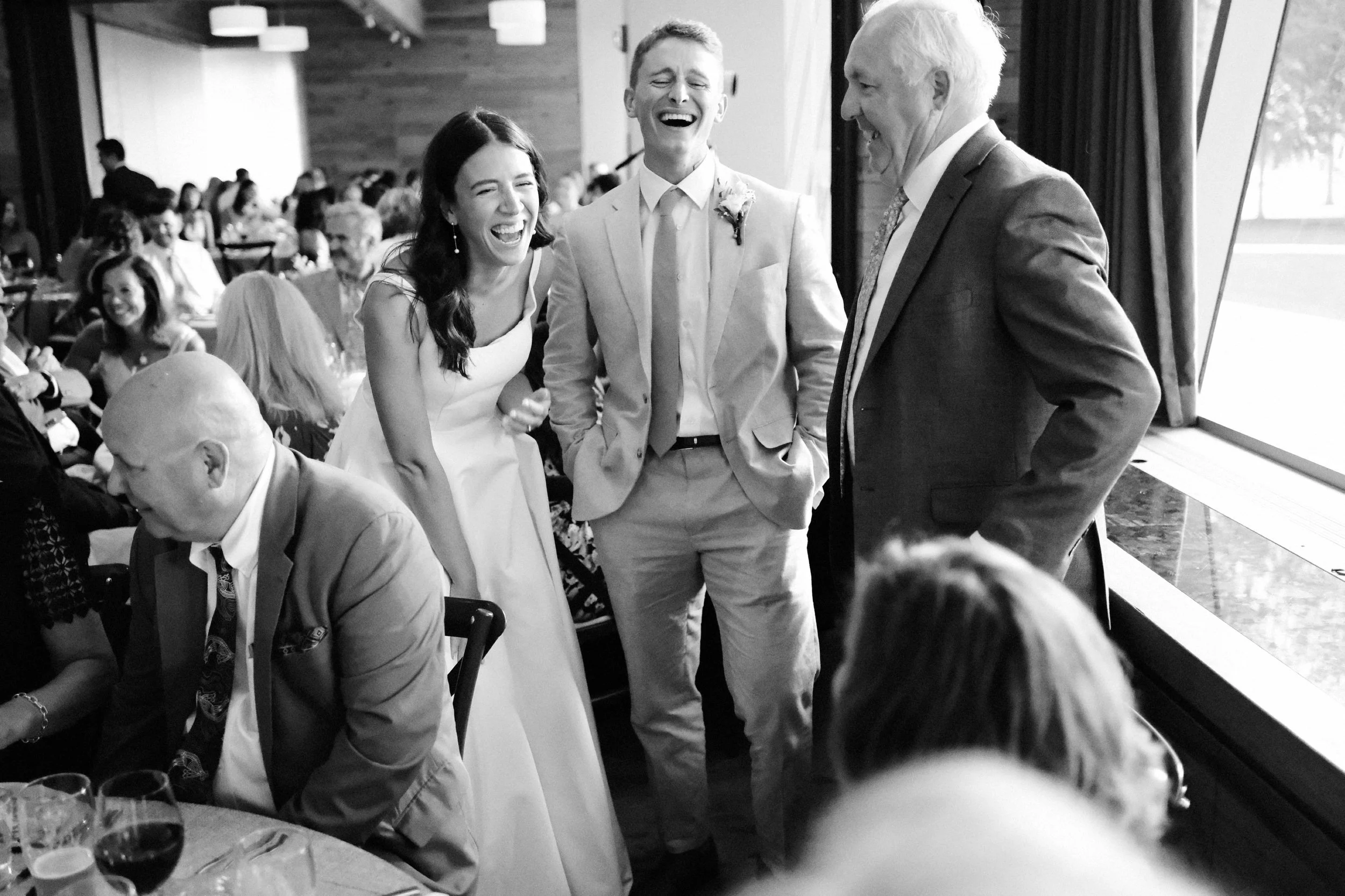 People laughing and socializing at a wedding reception, with a woman in a white dress, a man in a suit, and an older man in a suit near a window.