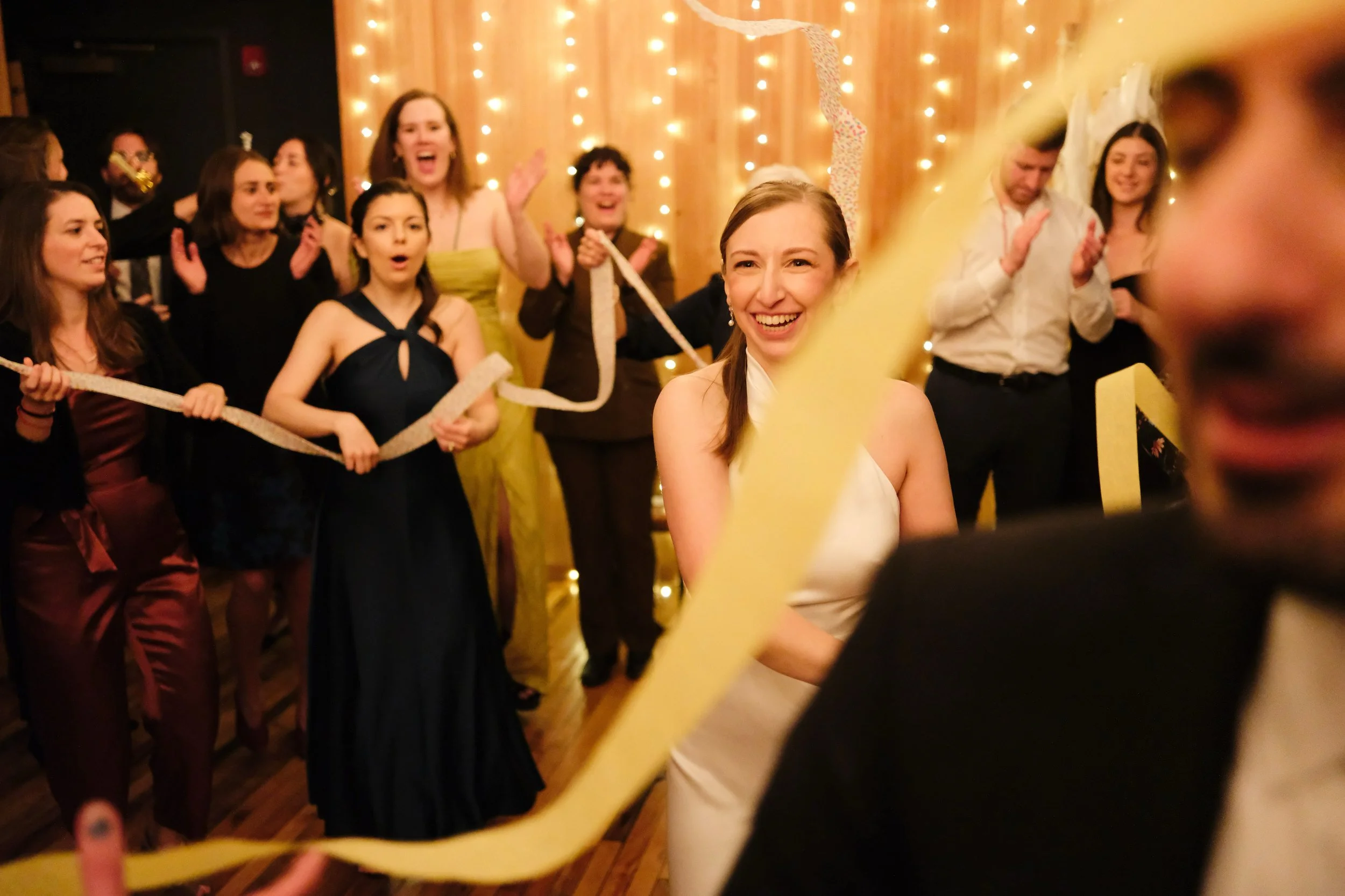 A group of people celebrating at a party with string and festive decorations, with a smiling woman in a white dress in the center.