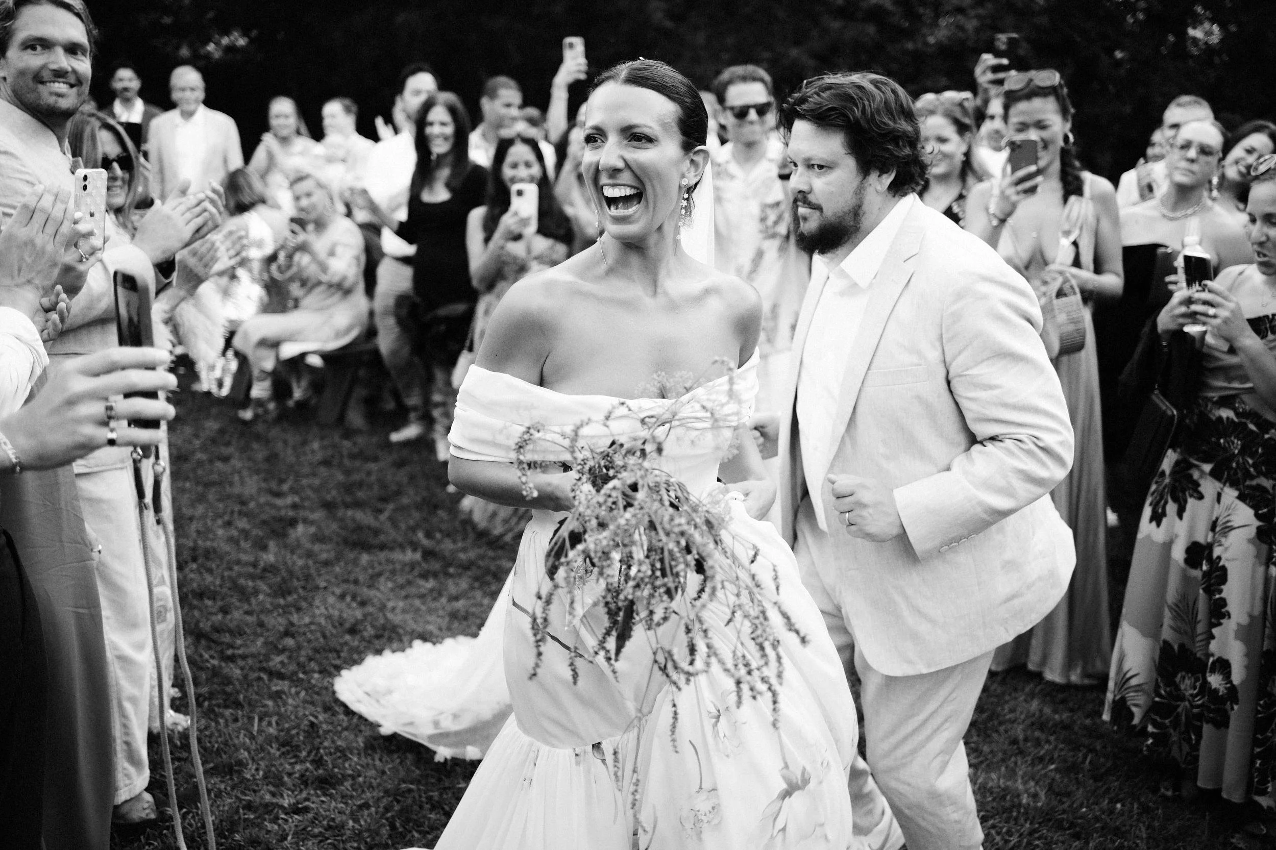 A black-and-white photo of a bride and groom smiling as they walk past a crowd of wedding guests. The bride is holding a bouquet and wearing an off-the-shoulder dress, while the groom is dressed in a light-colored suit.