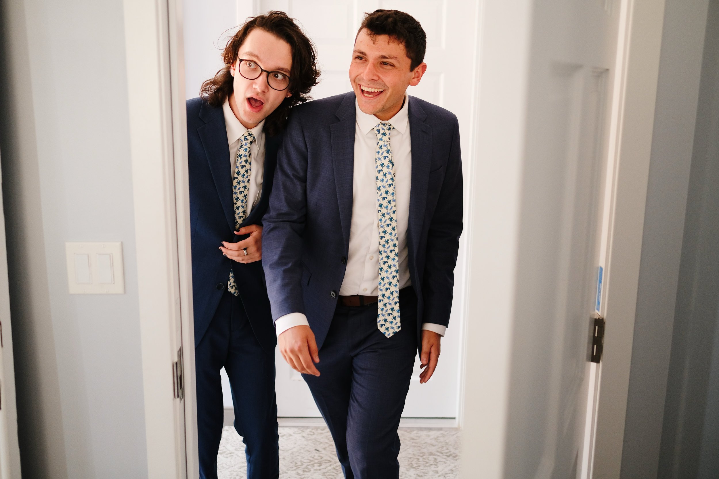 Two men in suits peek around a doorframe, both smiling and one with glasses, wearing coordination patterned ties.