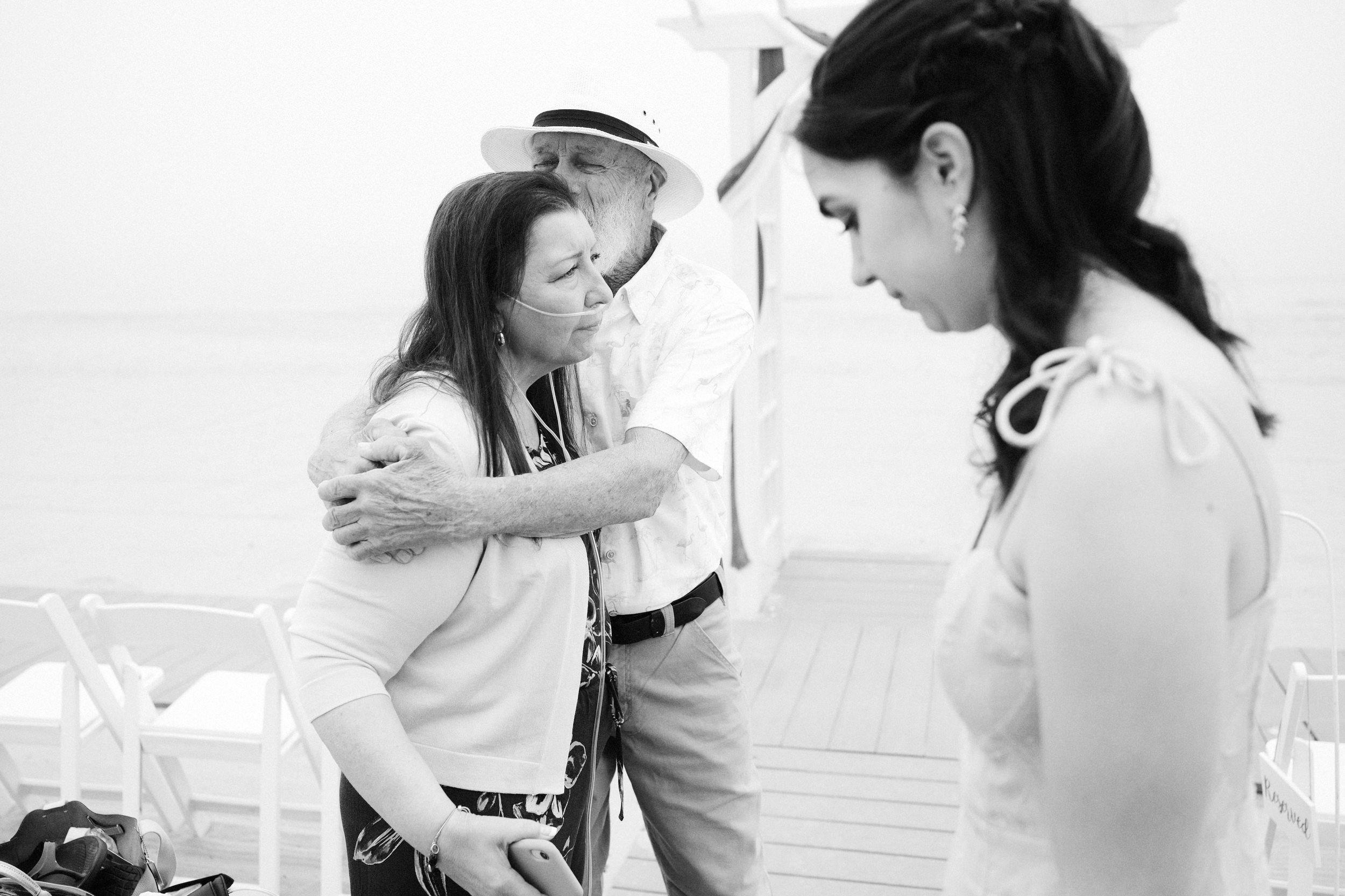 A woman receiving a hug from an older man while a nurse looks on during a wedding ceremony on a beach.
