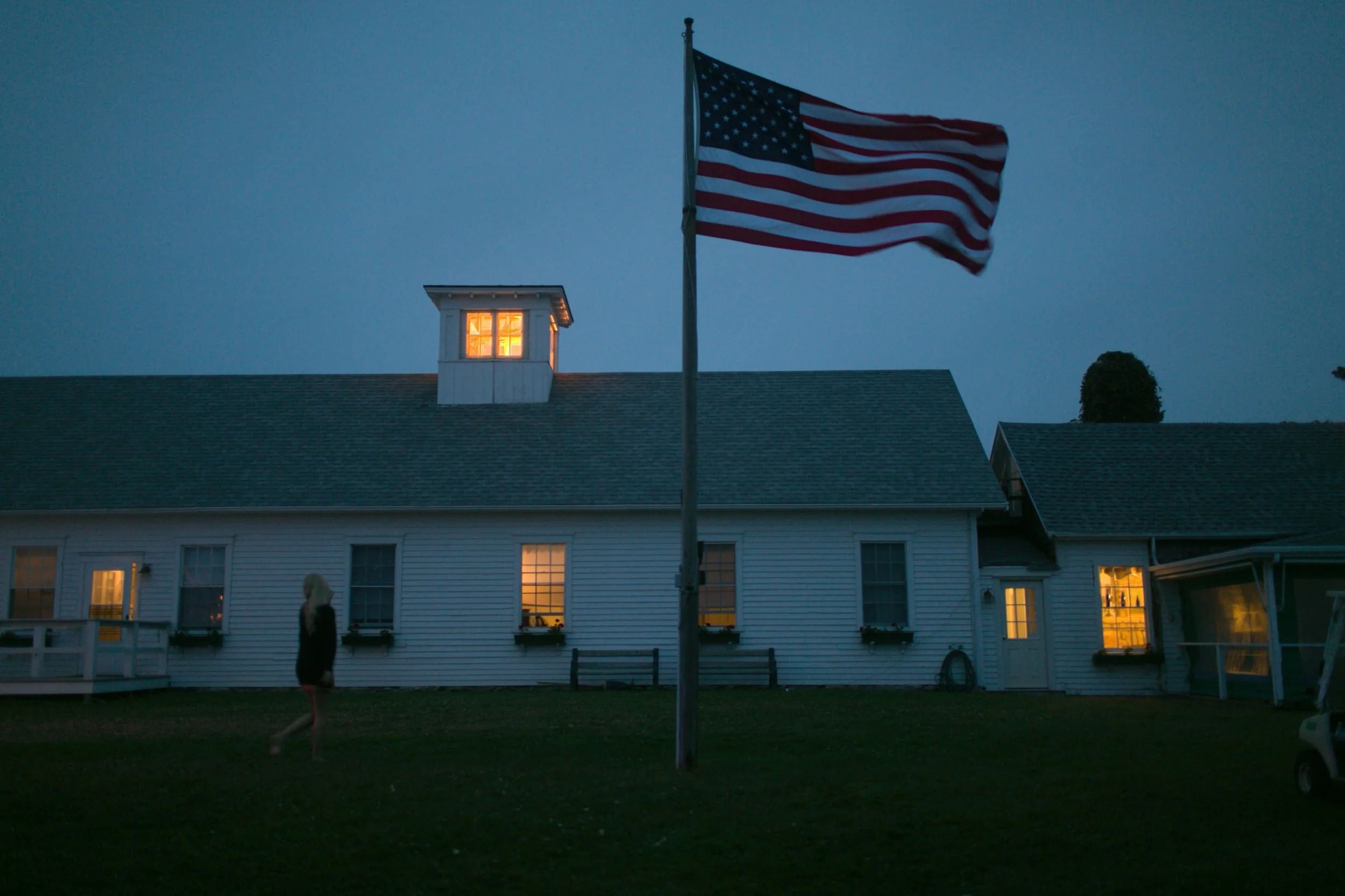 An American flag waving outside a white house at dusk, with warm light glowing from the windows.