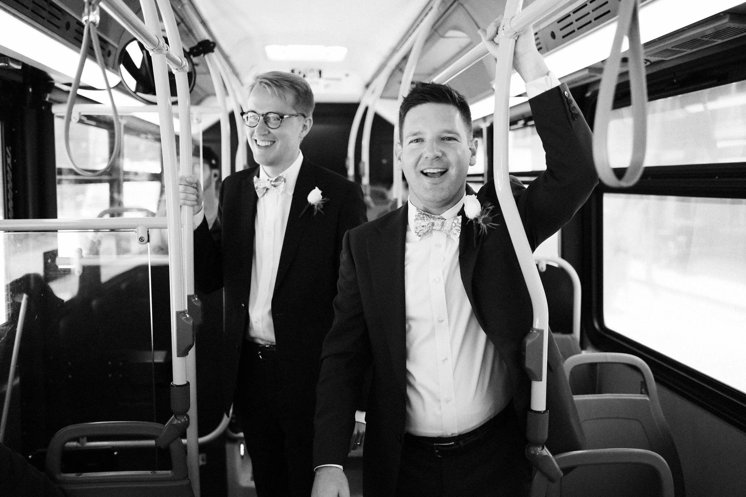 Two men in suits with bow ties and boutonnières on a bus, smiling and holding onto the bus handrails.