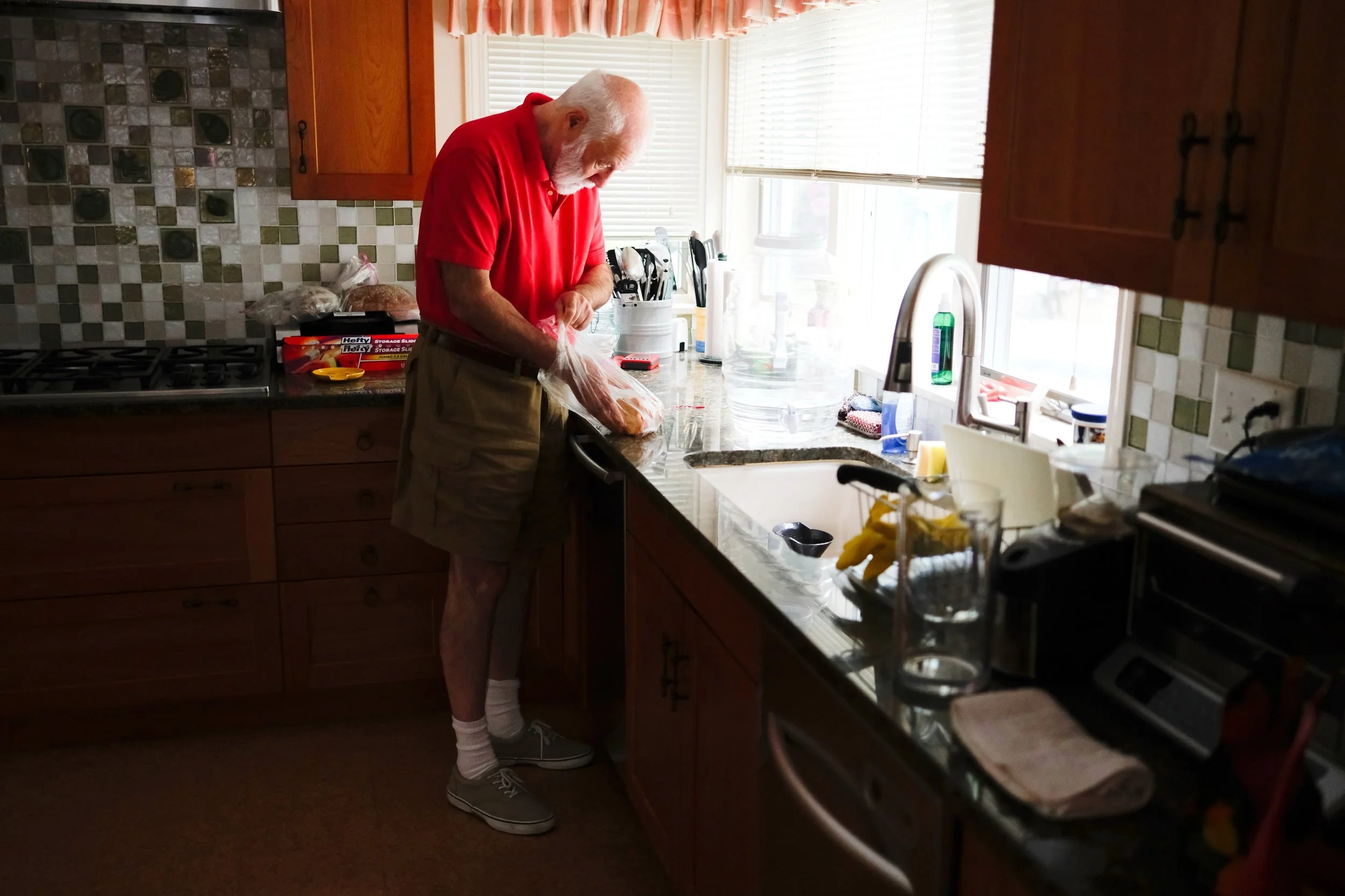 An elderly man in a red shirt and beige shorts stands in a kitchen, preparing food with a plastic bag on the counter near a window.