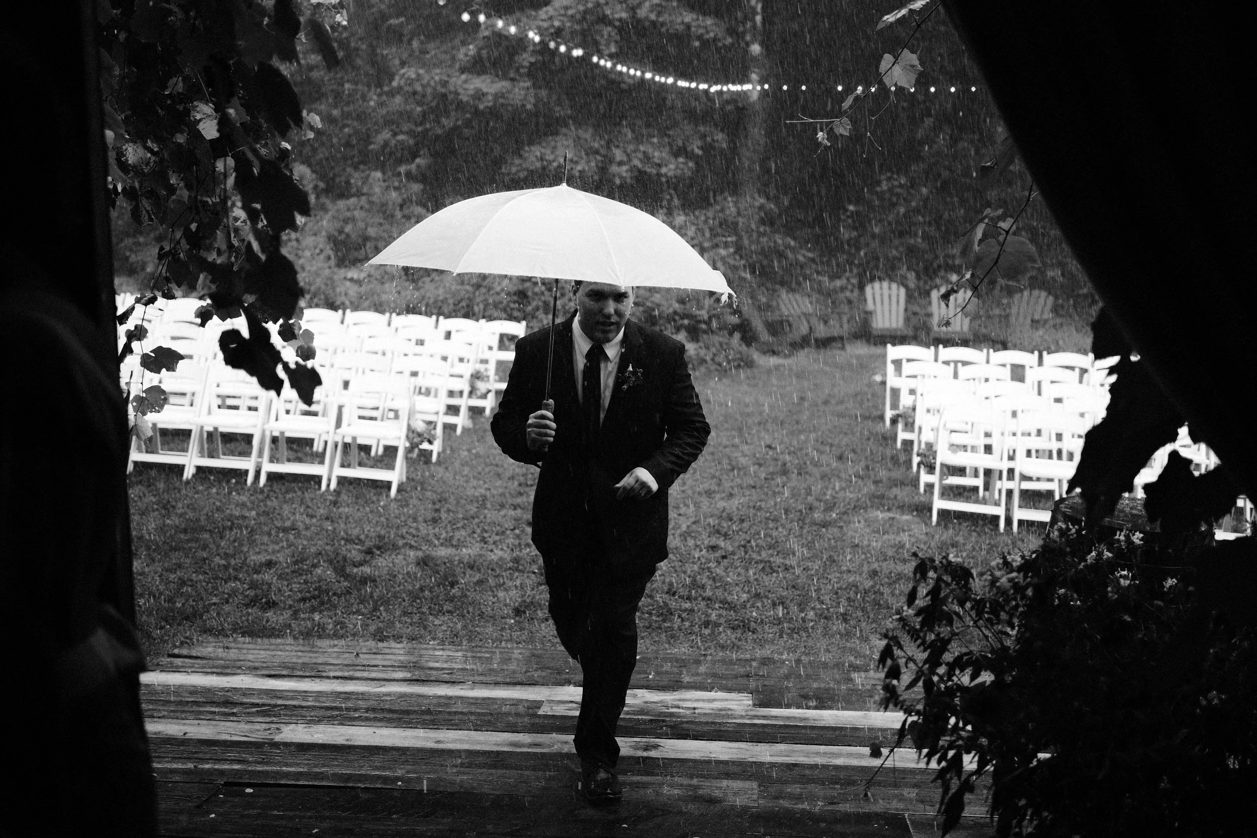 A man in a suit holding an umbrella walks through the rain on an outdoor wedding ceremony site with rows of white chairs, surrounded by trees and foliage.