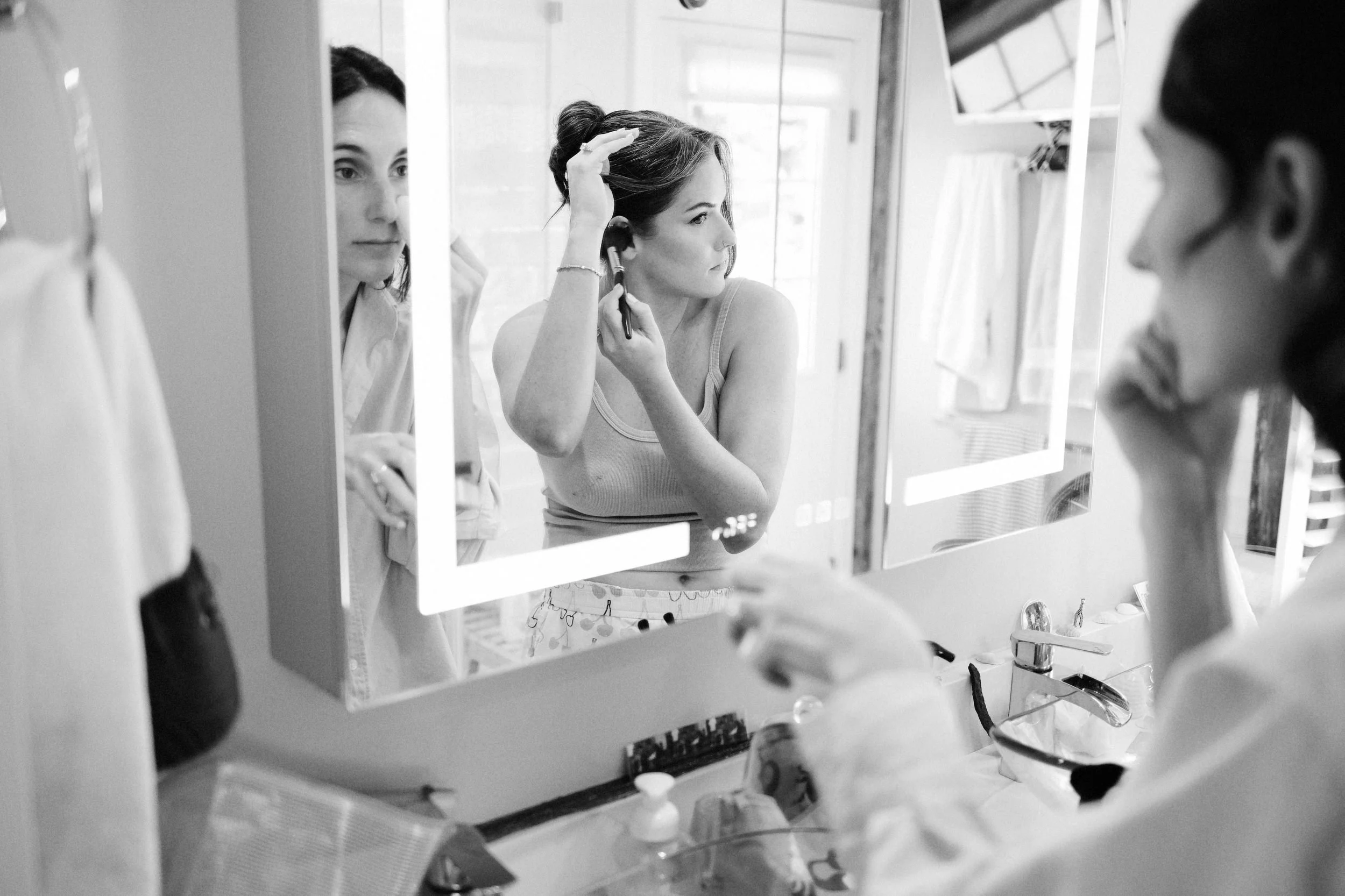 A woman looks at her reflection in a bathroom mirror, applying makeup, with another woman standing nearby.