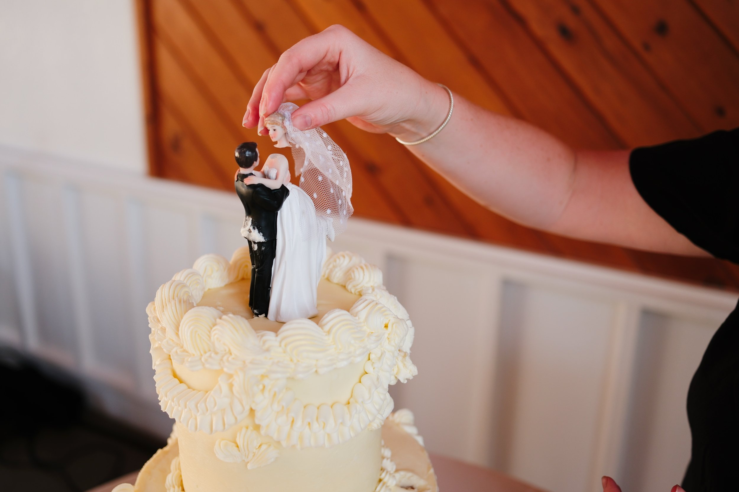 A wedding cake topper featuring a bride and groom figurine, with a woman's hand adjusting the bride figure's veil.