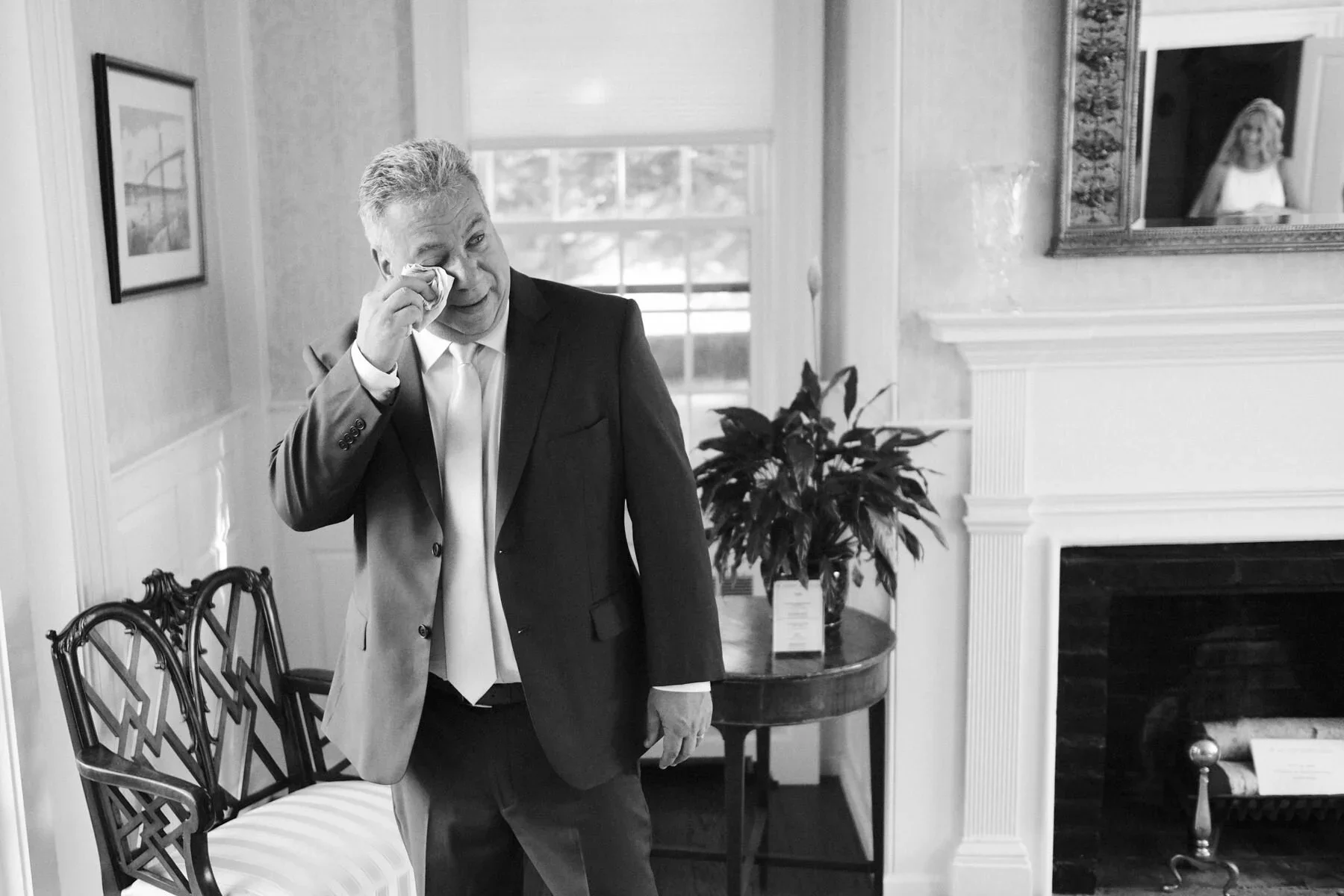 A man in a suit, wiping his eyes with a tissue, standing in a room with a fireplace, a table with a plant, and a framed picture on the wall, with a woman looking at him through a mirror.