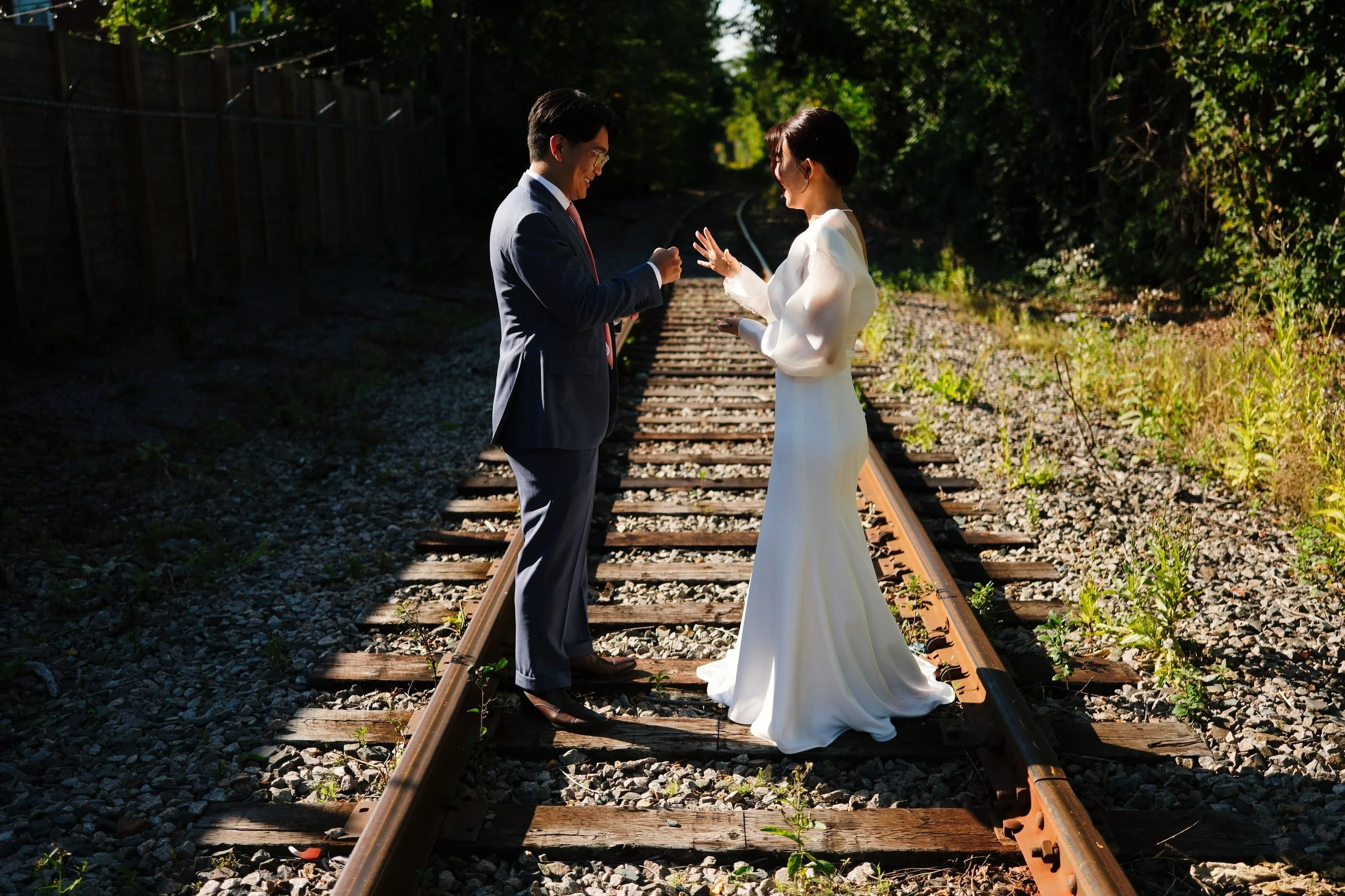 A bride and groom standing on railroad tracks outdoors, facing each other, with the groom in a dark suit and the bride in a white wedding dress, illuminated by sunlight.