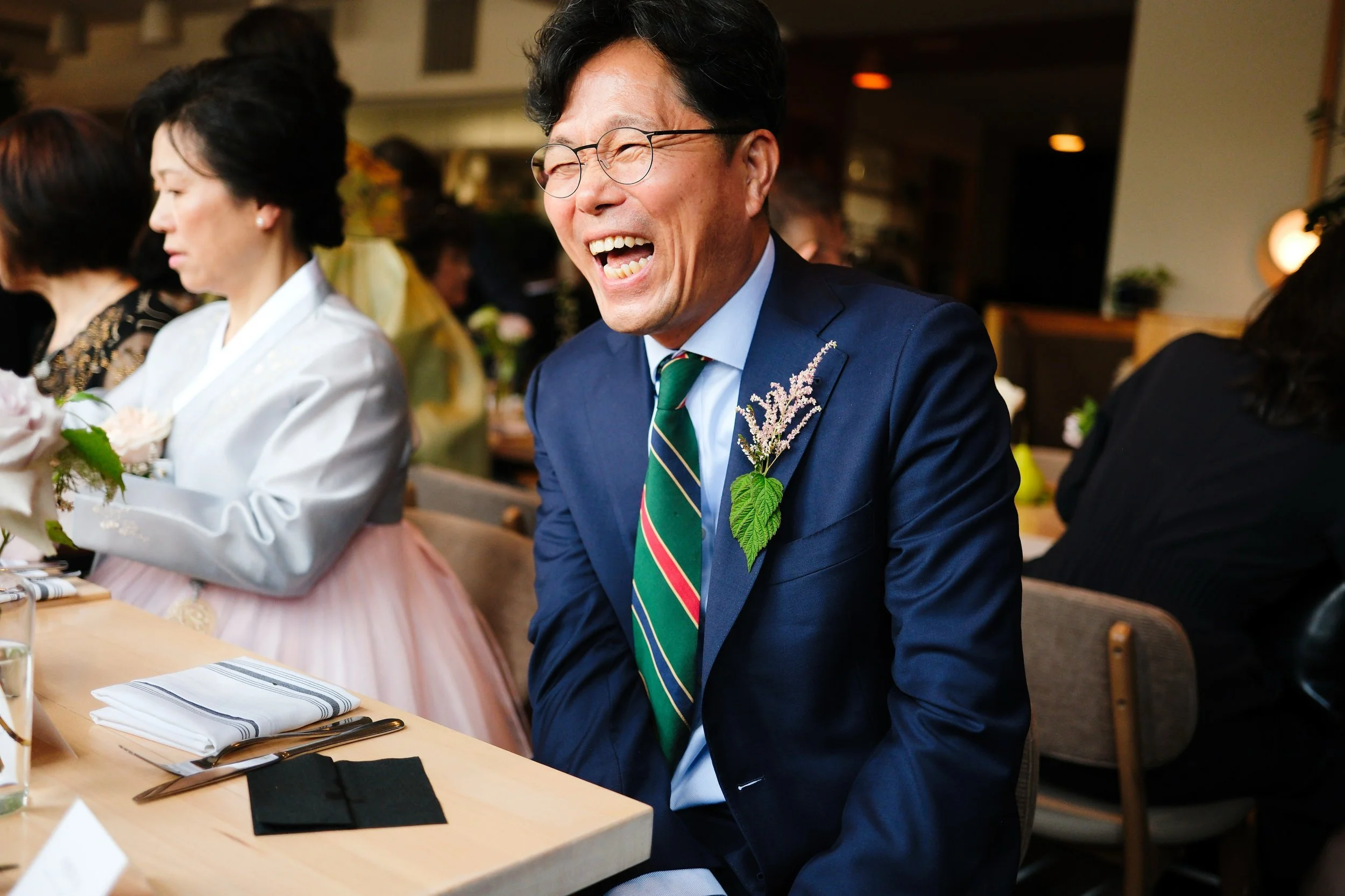 A man in a navy suit and striped tie, wearing glasses, sits at a table laughing during a celebration. He has a sprig of flowers and leaves pinned to his jacket. Other people are sitting at the same table, dressed nicely, in a warmly lit indoor settin