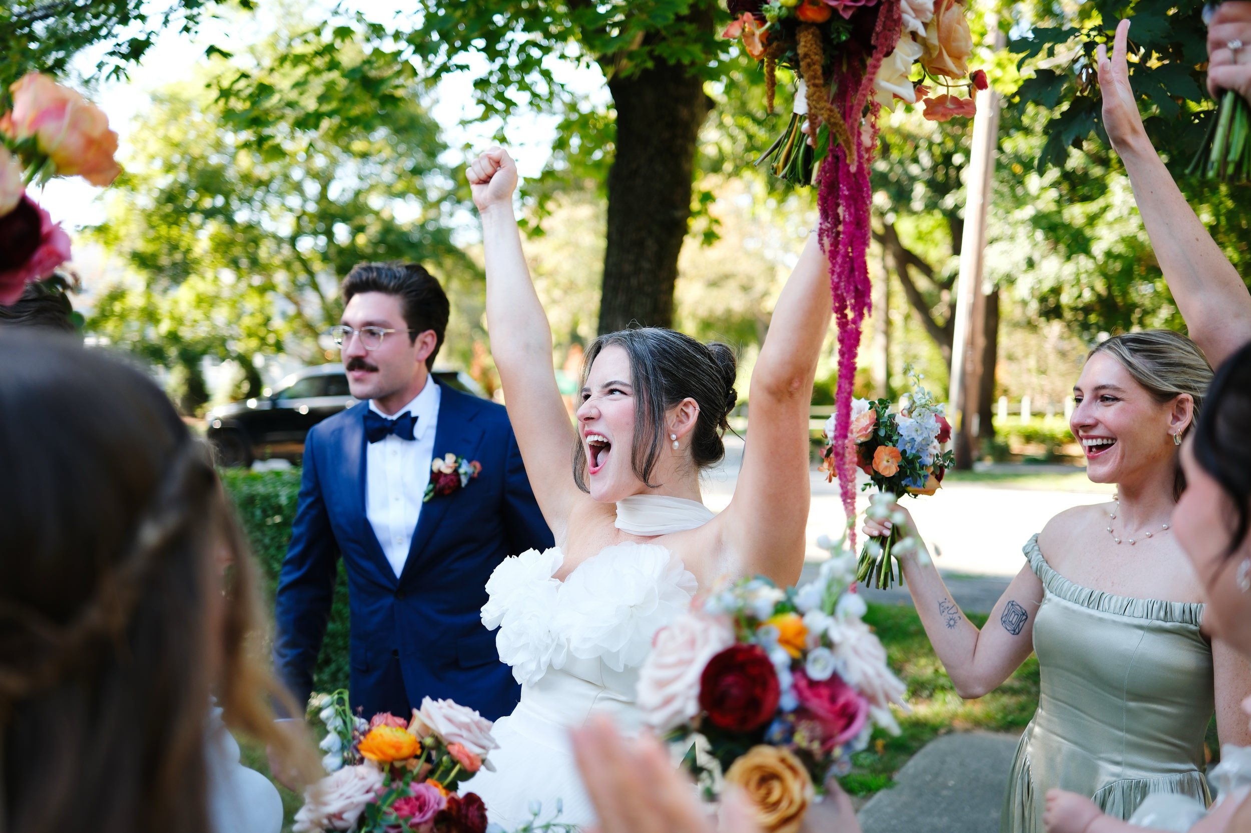 A woman in a white wedding dress celebrating with her arms raised, surrounded by wedding guests outdoors on a sunny day, with flowers and greenery in the background.