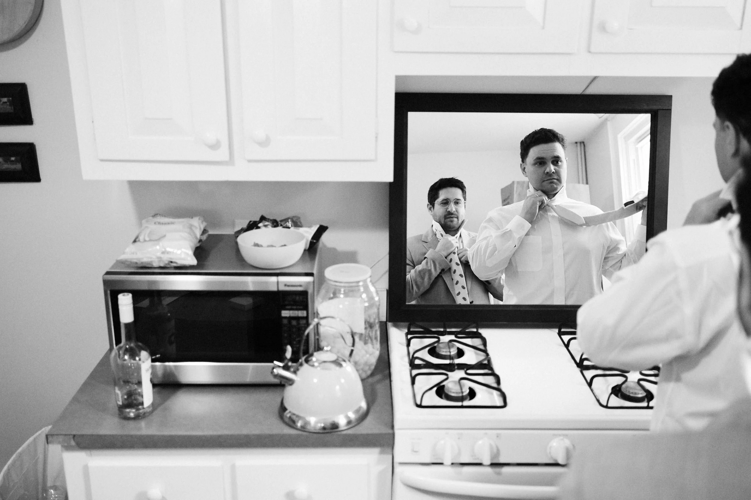 A man looks in a kitchen mirror as another man with a beard adjusts his tie, both dressed in suits. The reflection shows they are getting ready, one tying his necktie and the other adjusting his bowtie.