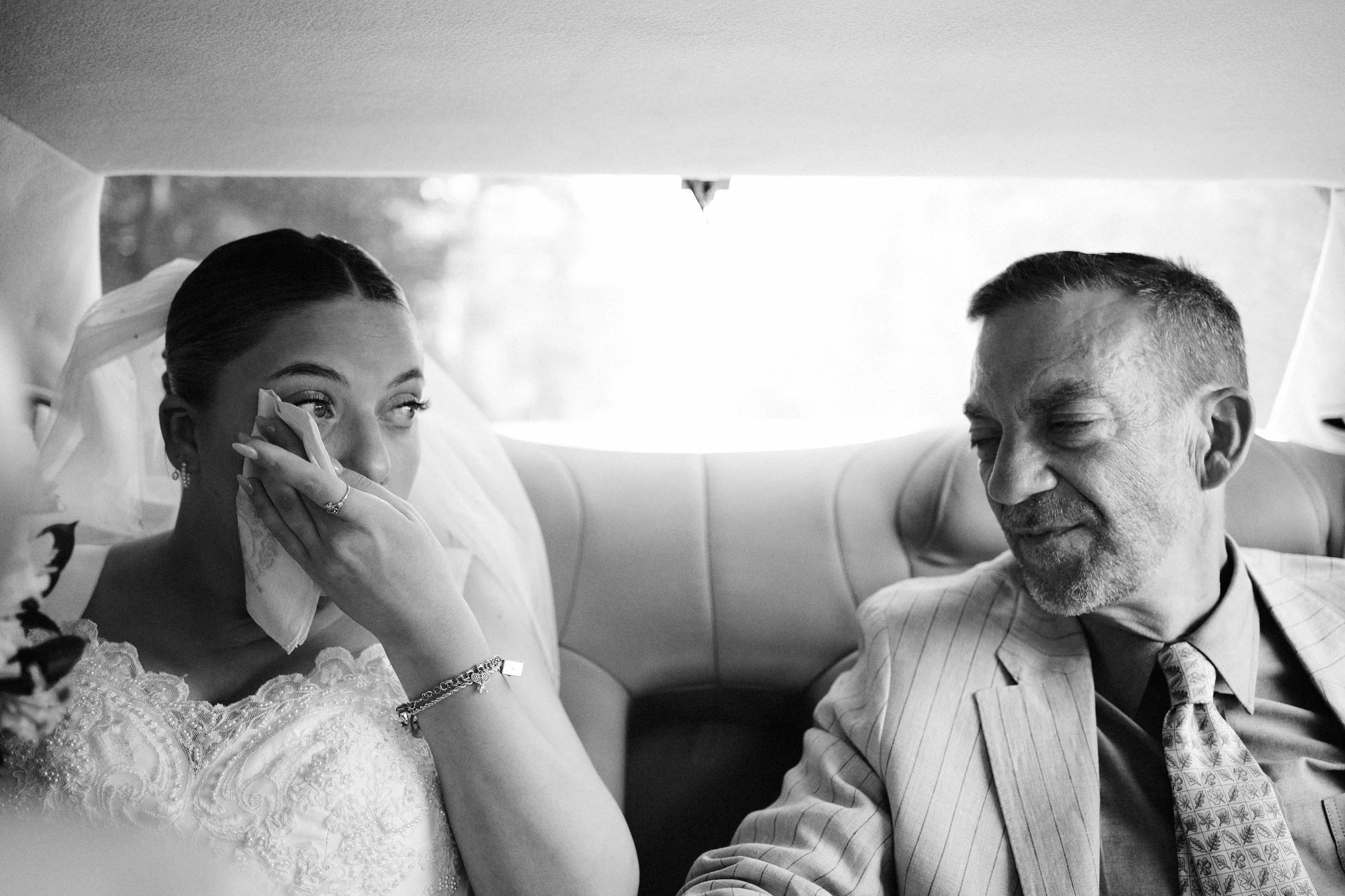 A bride wiping her tears while sitting in a vehicle with an older man, possibly her father, during a wedding moment in black and white.