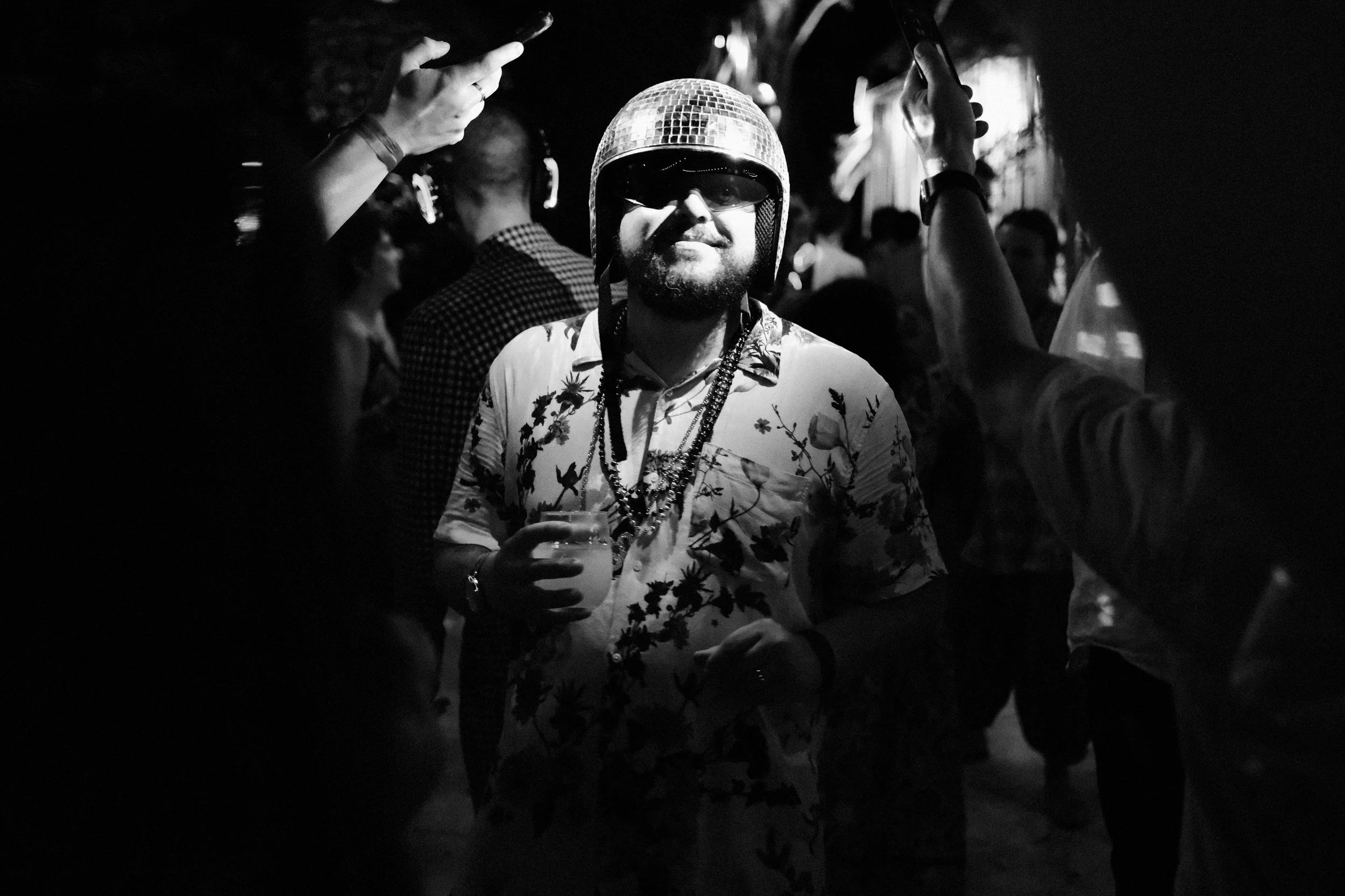 A man wearing a motorcycle helmet and sunglasses at a party, holding a drink, and surrounded by other people, some with their phones raised.