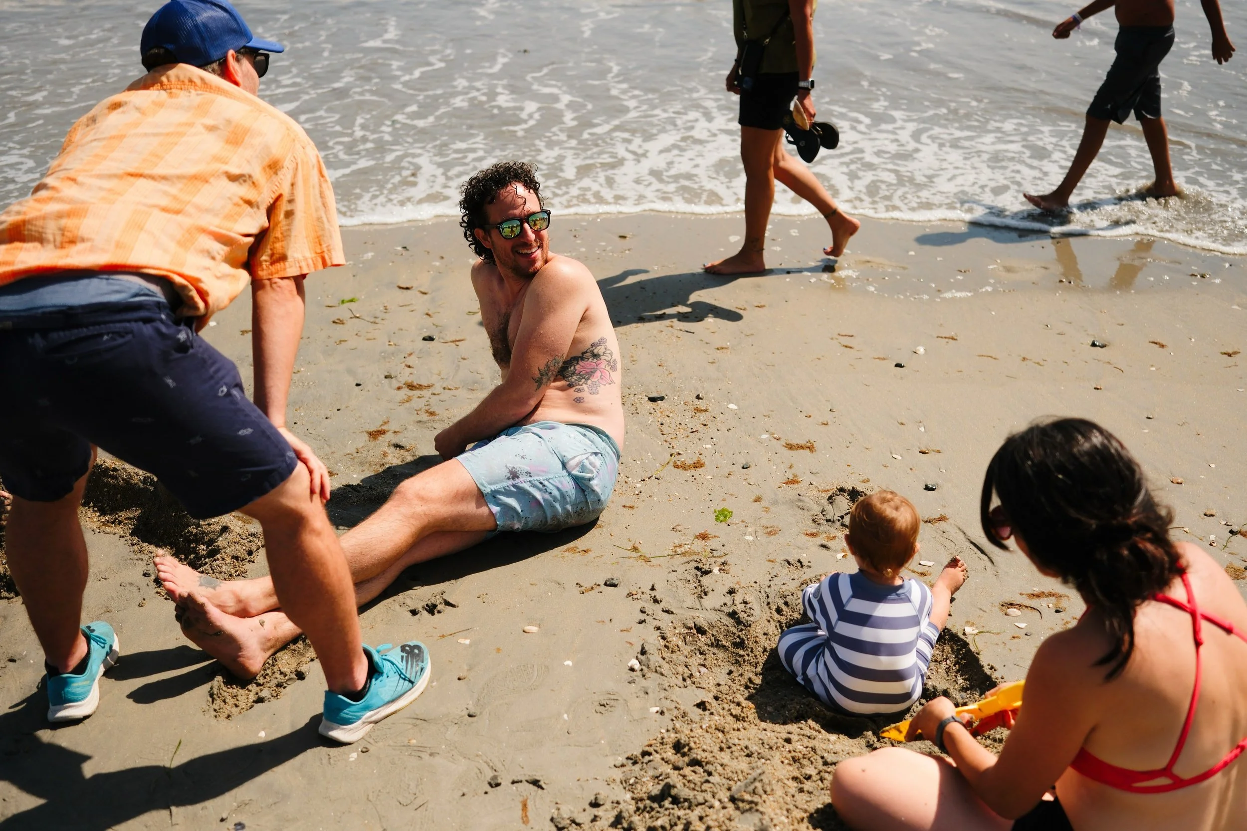 People at the beach, with one man sitting in the sand, smiling and talking to another man kneeling beside him. Two children are playing nearby, one digging in the sand. Others are walking in the water.