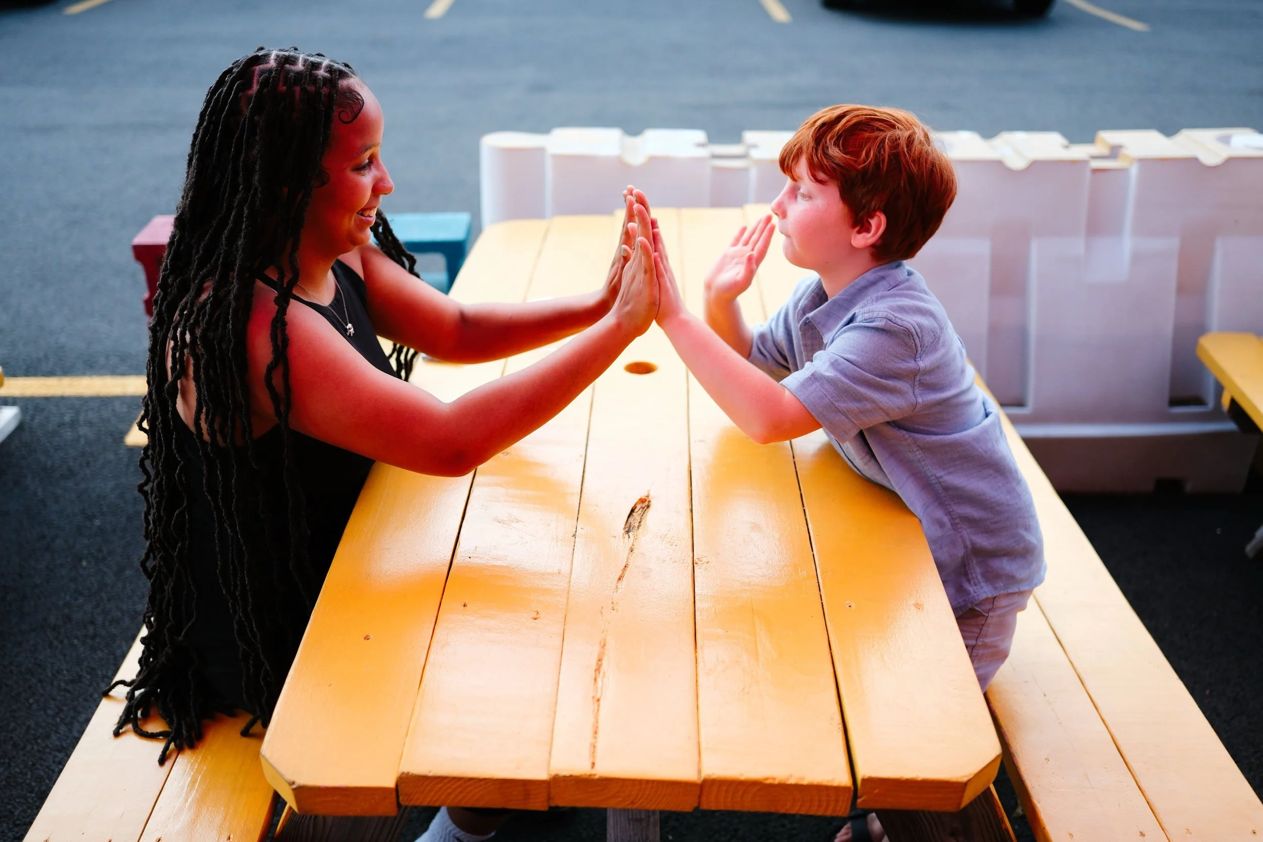 Two children, a girl with long black dreadlocks and a boy with red hair, are engaging in a friendly game of wall ball while sitting at an outdoor wooden picnic table. They are facing each other, with palms pressed together, smiling and playing on a c