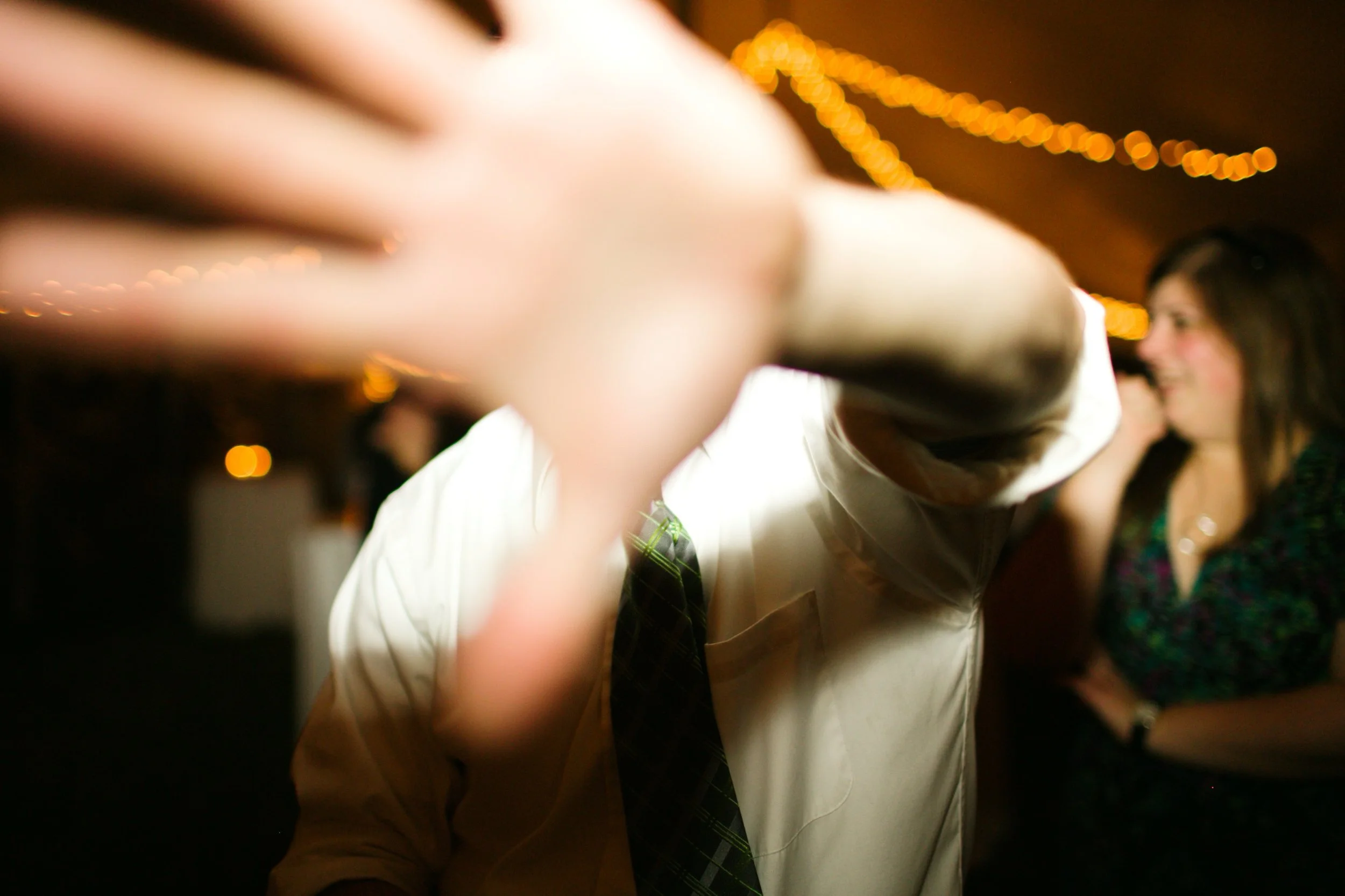 A person with a blurred hand reaching towards the camera, with a woman smiling and talking in the background at a party or social gathering under warm string lights.