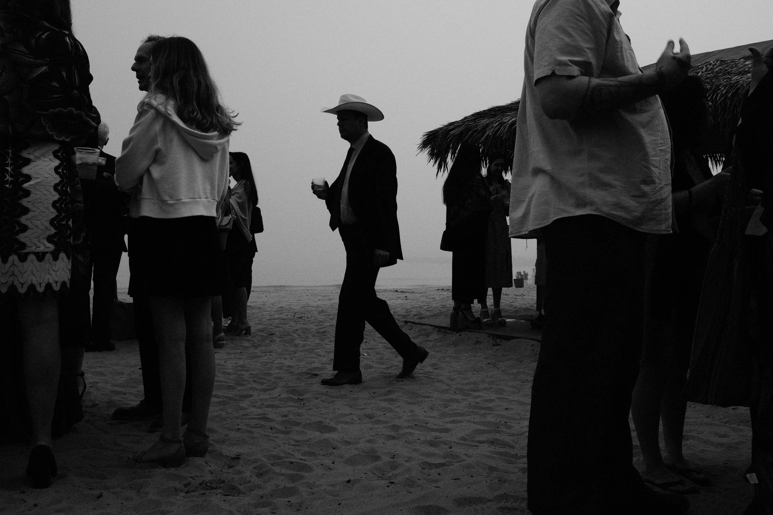 People gathering on the beach, some holding drinks, with a man wearing a cowboy hat in the center, and a thatched-roof hut in the background, black and white photo.