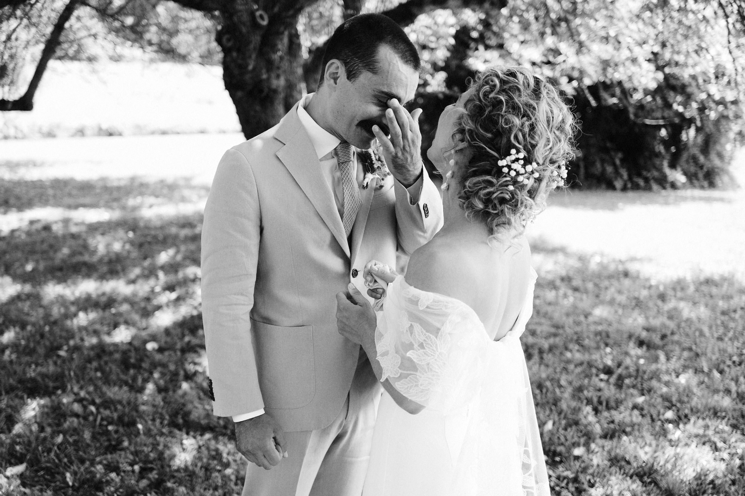 Black and white photo of a bride and groom outdoors under a tree, sharing a joyful moment with the groom touching his face and the bride holding a ring.