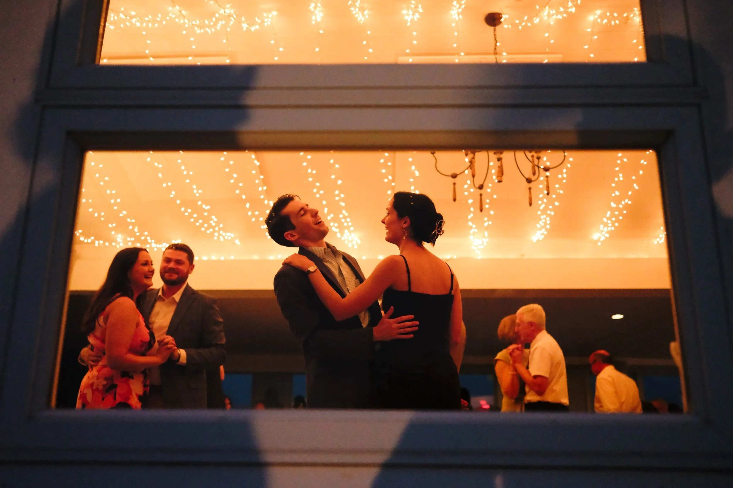 People dancing and socializing at a wedding reception viewed through a window, with string lights and a chandelier illuminating the room.