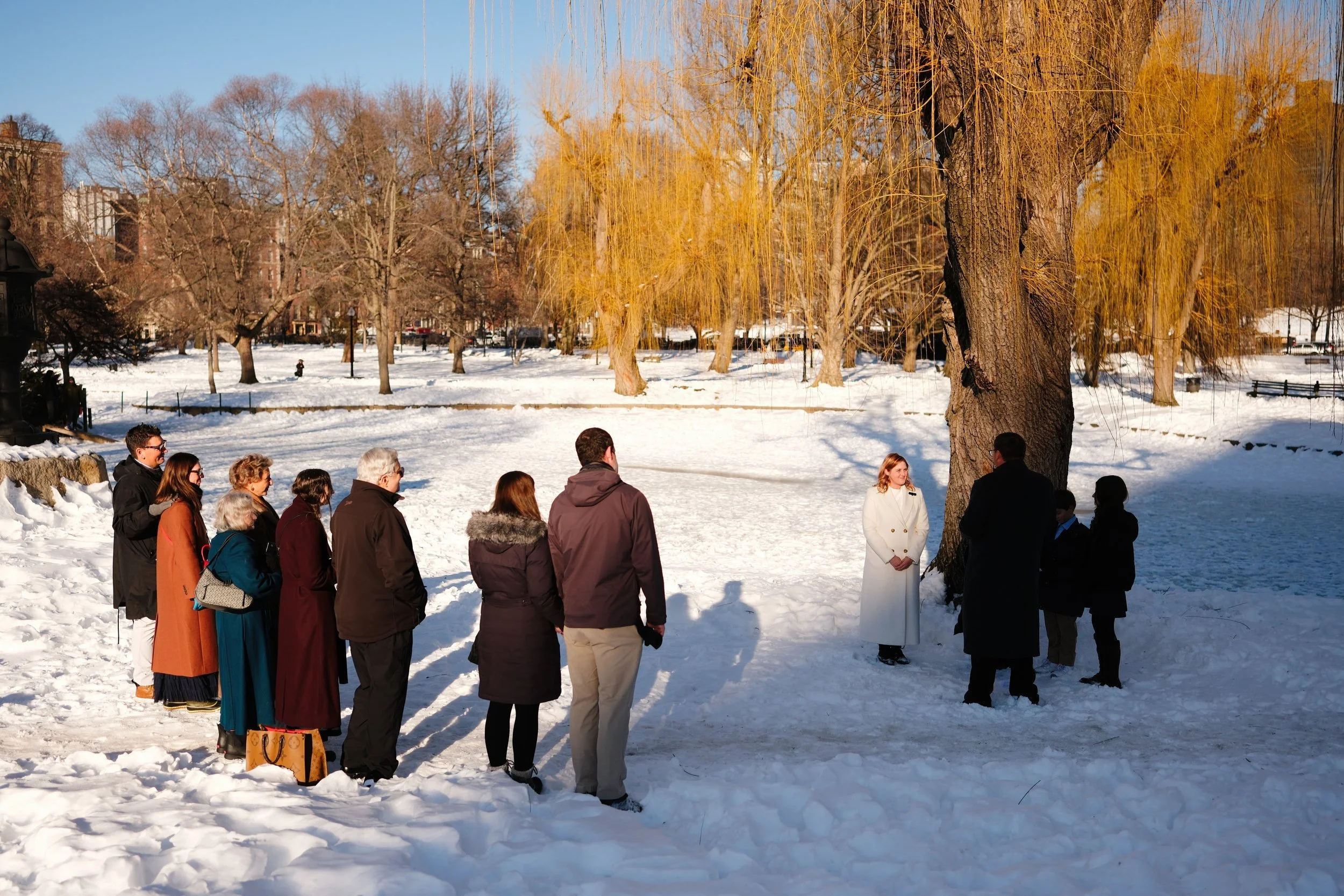A group of people gathered outdoors in a snowy park, attending a ceremony or event under a large tree, with some participants dressed warmly and a woman dressed in white standing apart from the group.