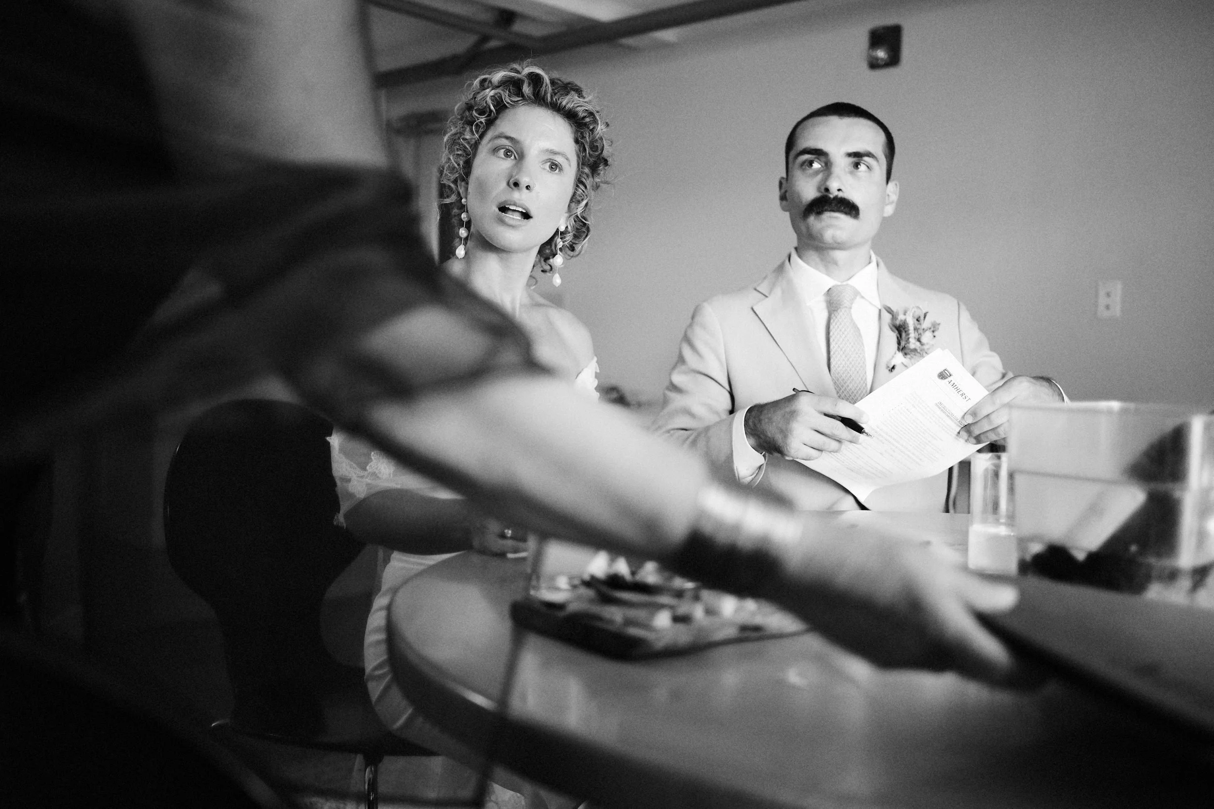 A black and white photo of a woman with curly hair and earrings, and a man with a mustache in formal attire, seated at a table with documents. An arm and hand are visible in the foreground.