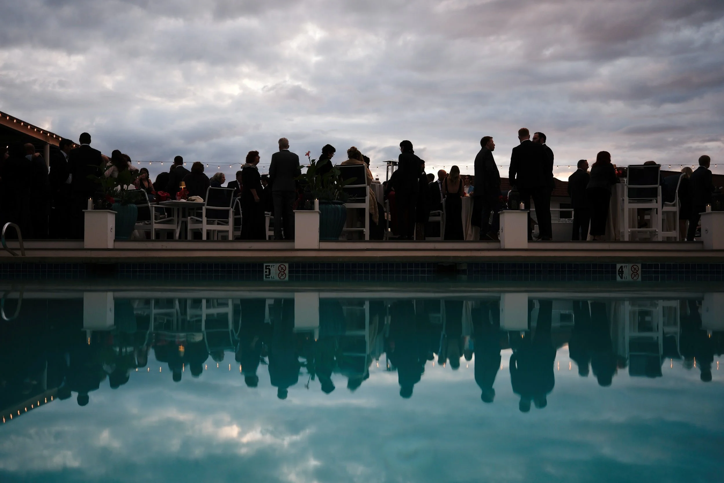 Group of people in formal attire gathered on a rooftop terrace around tables, with a swimming pool reflecting the cloudy evening sky in the foreground.