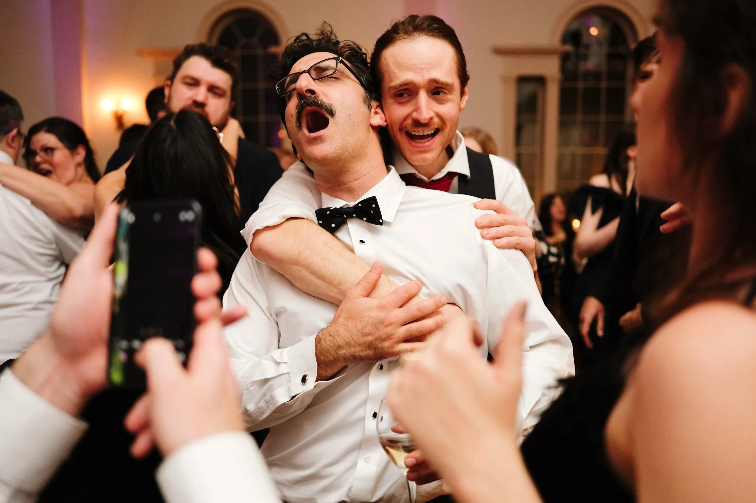 People at a party or celebration dancing and hugging, with one man in a tuxedo, glasses, and a bow tie appearing to sing or shout with a woman in a black dress showering him with affection, other guests enjoying the moment in the background.