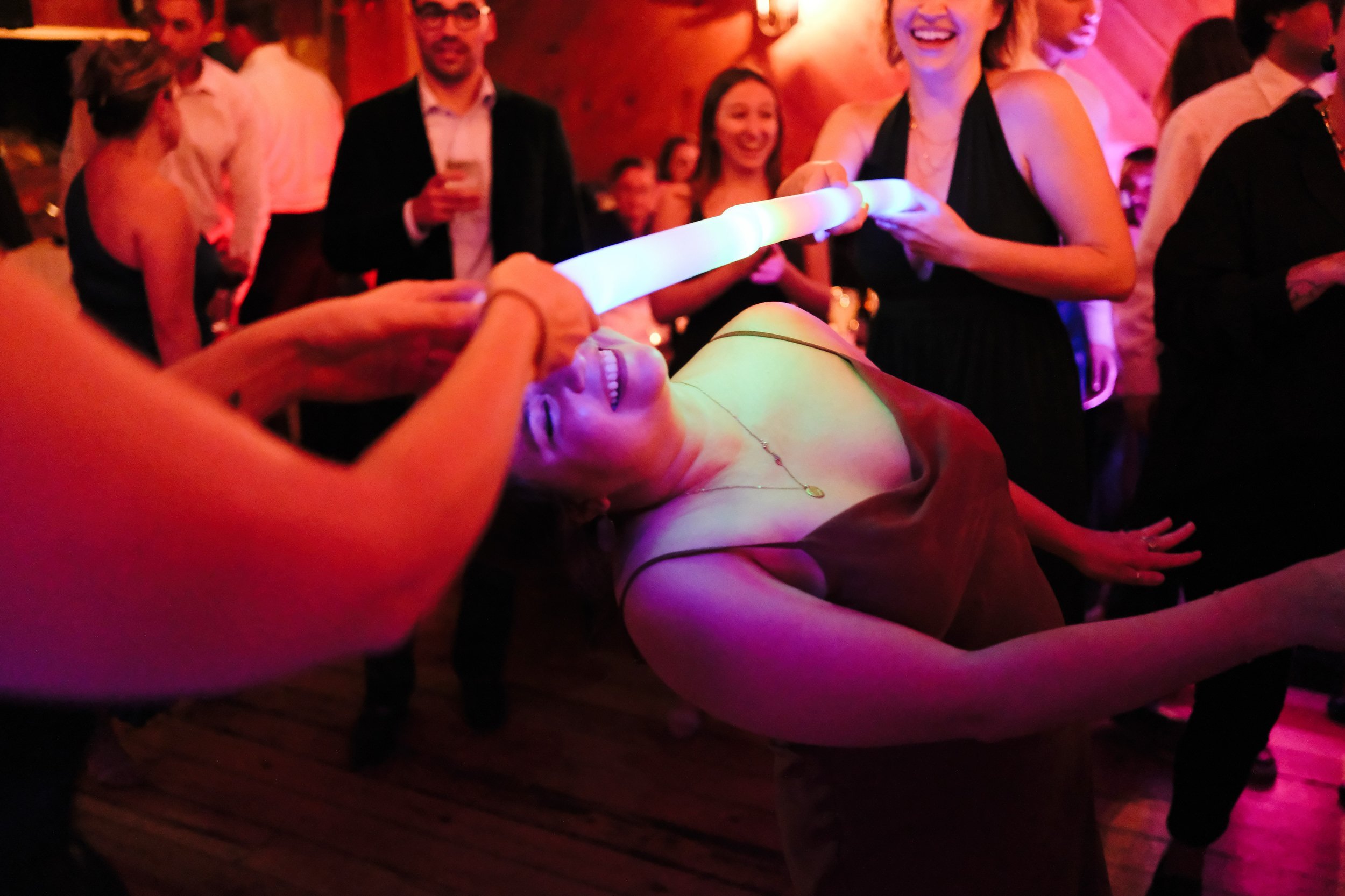 People at a party with one woman lying back while another person feeds her a glowing, illuminated object, with other guests smiling and chatting in the background.