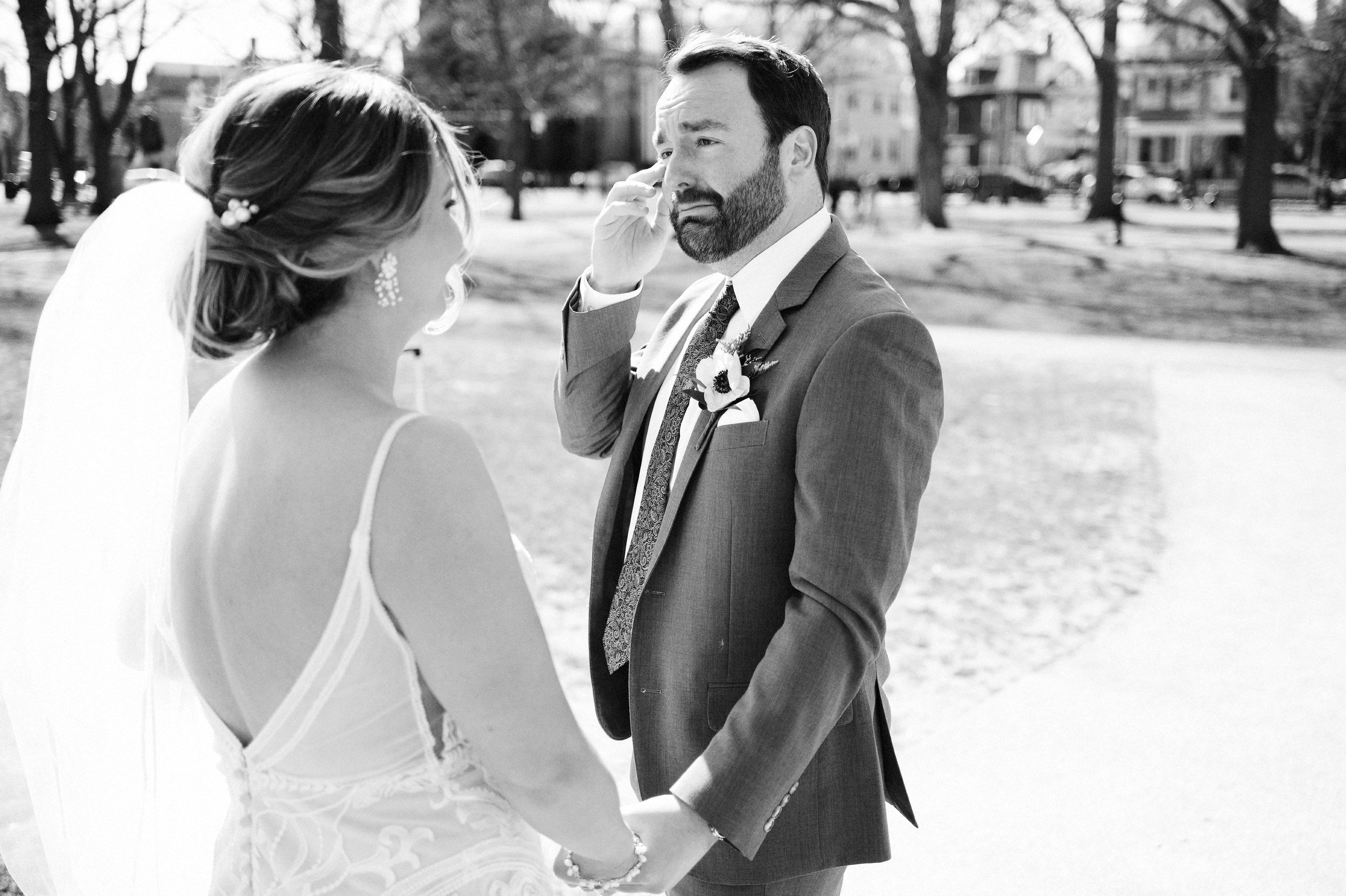 A bride and groom holding hands during their outdoor wedding ceremony in a park, with trees and houses in the background.