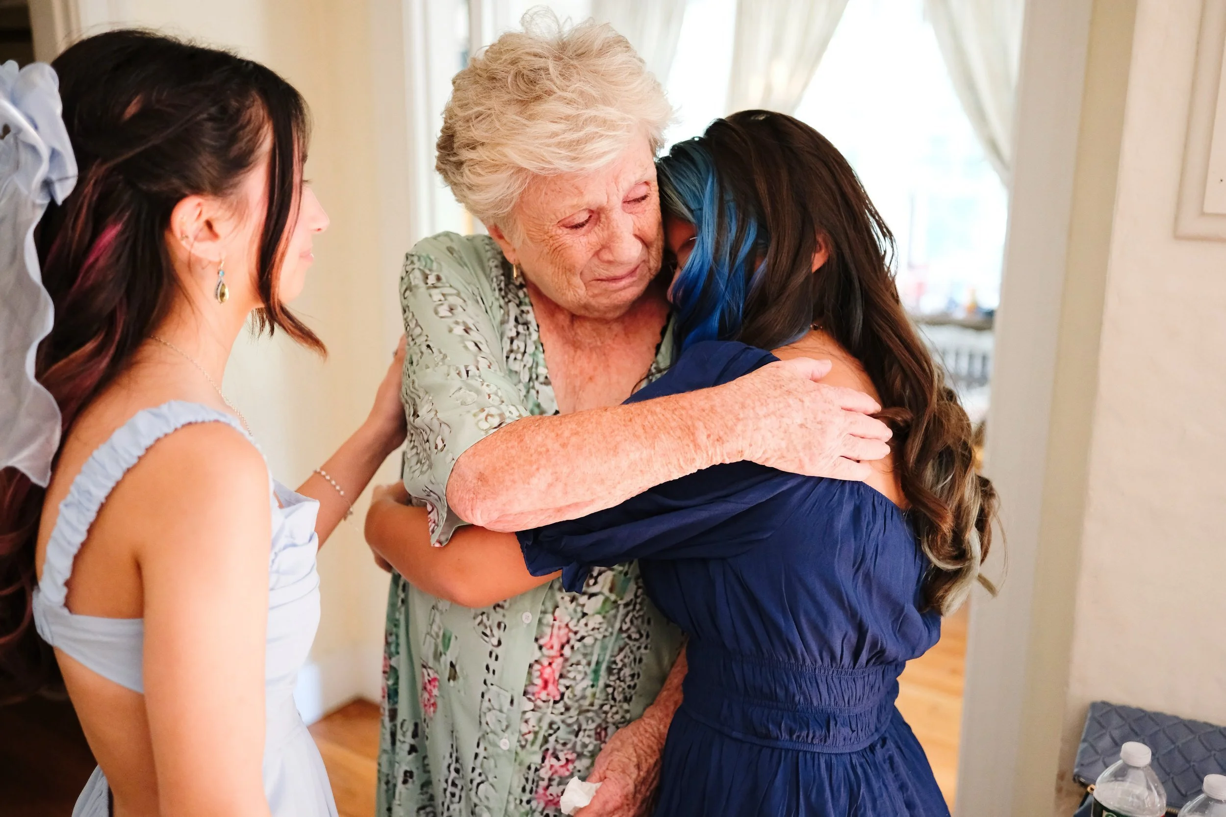 An elderly woman hugging a young woman with blue streaks in her hair, surrounded by other people, in a warmly lit room.