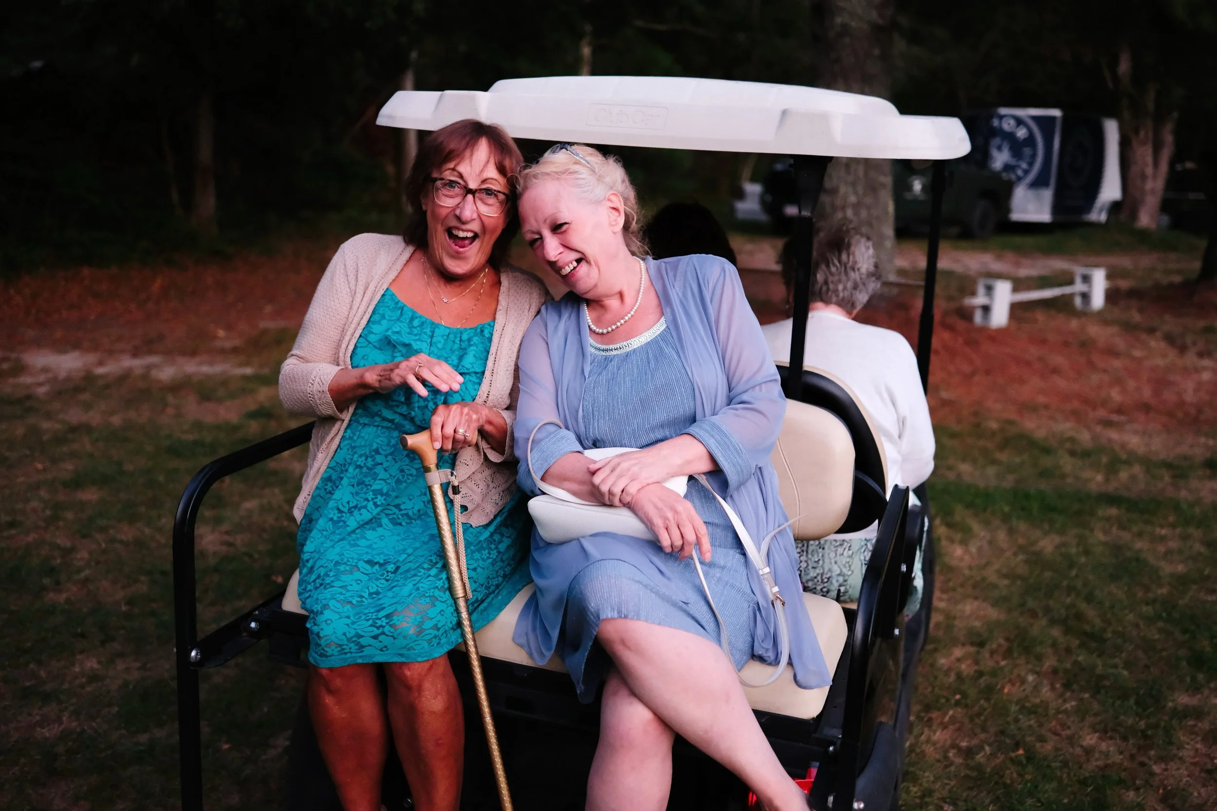Two elderly women laughing and sharing a joyful moment on a golf cart outdoors, with one woman holding a cane.