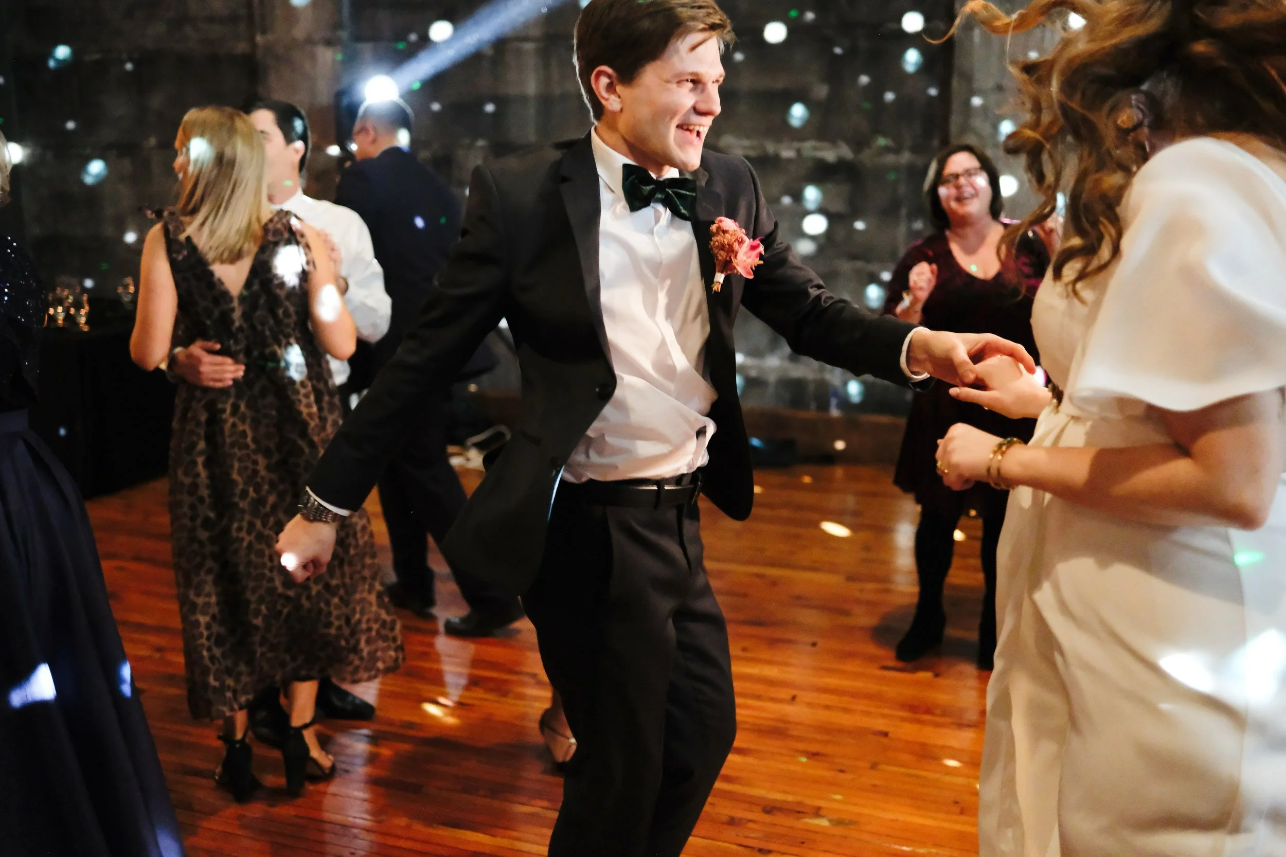 People dancing and socializing at a party, with a man in a tuxedo and a woman in a white dress holding hands in the foreground, and others in elegant attire in the background.