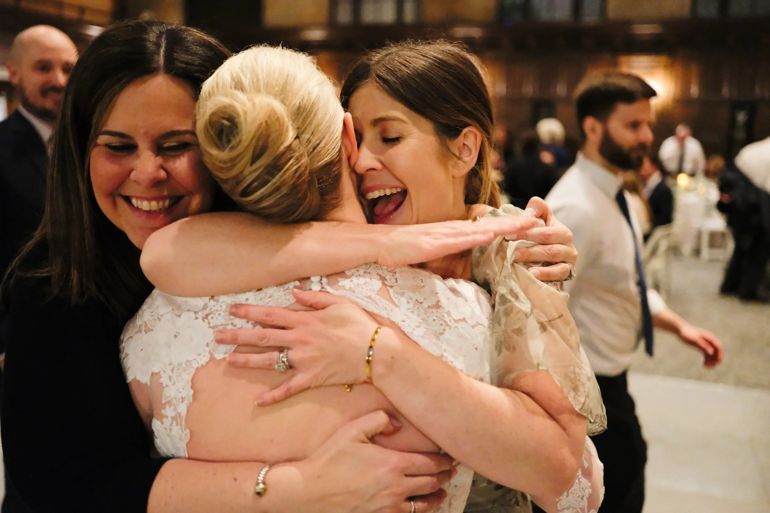 Three women hugging and smiling at a wedding celebration. In the background, there are other guests in a warmly lit, rustic venue.