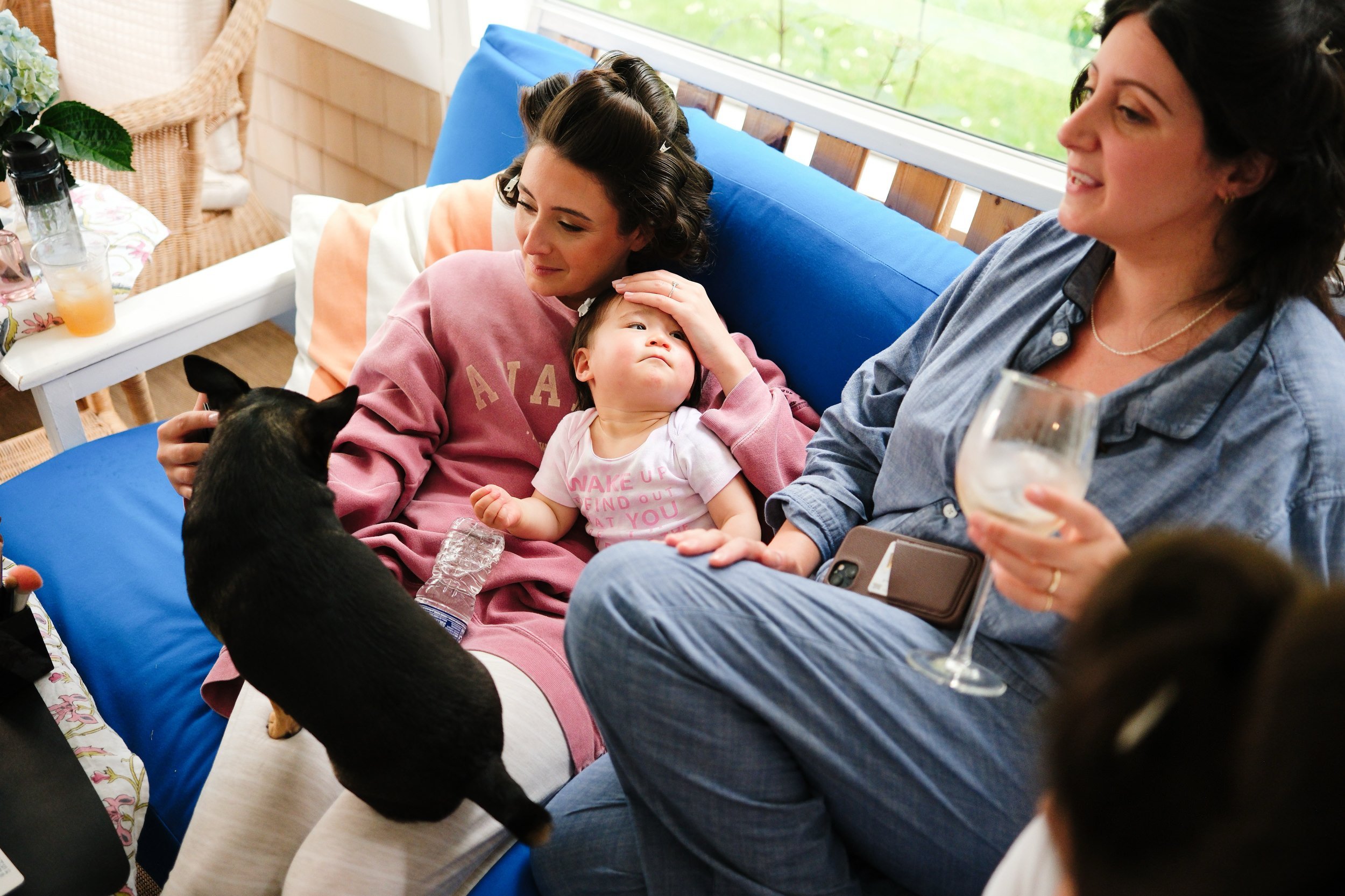 Two women, a young child, and a small black dog sitting on a blue sofa in a sunlit room. One woman is touching the child's head, and the other woman is holding a wine glass, smiling.