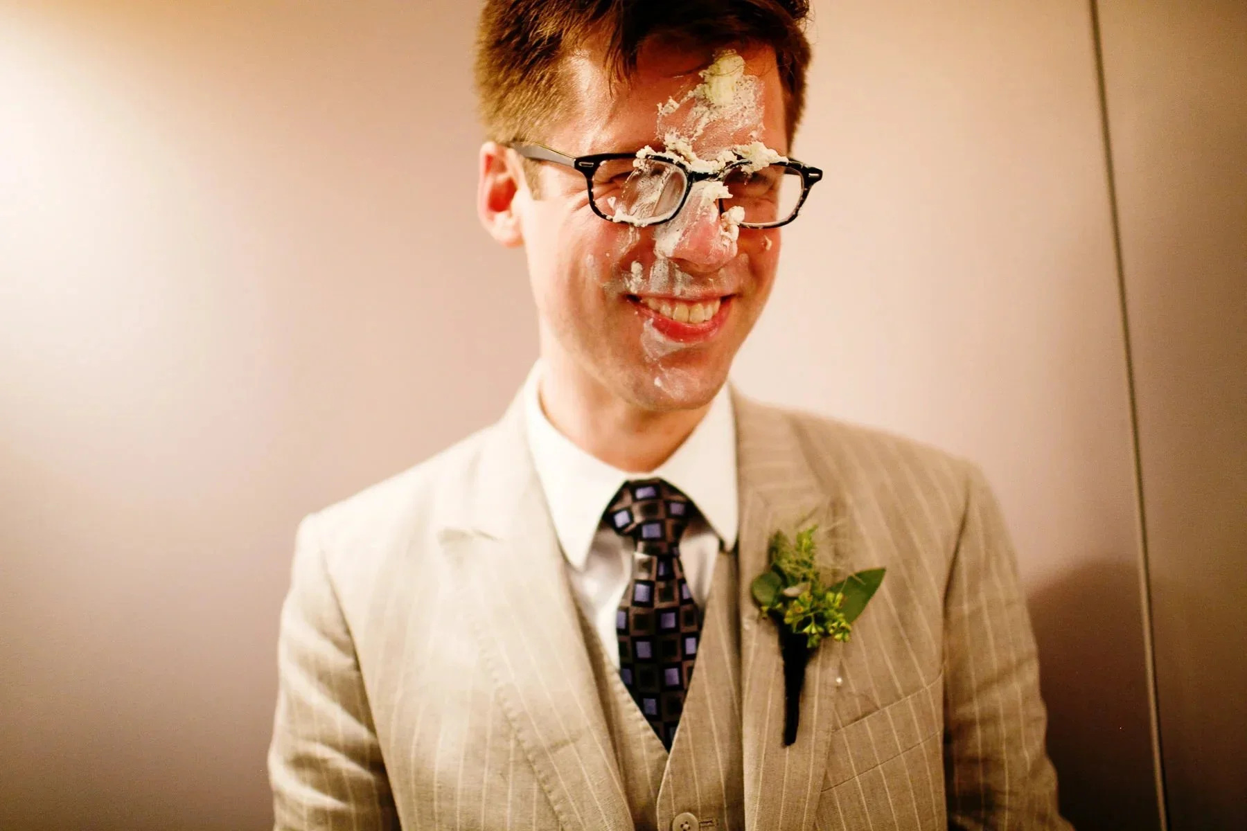 A man in a beige suit with a boutonniere, wearing glasses, has whipped cream and cake on his face and glasses, smiling in an elevator.